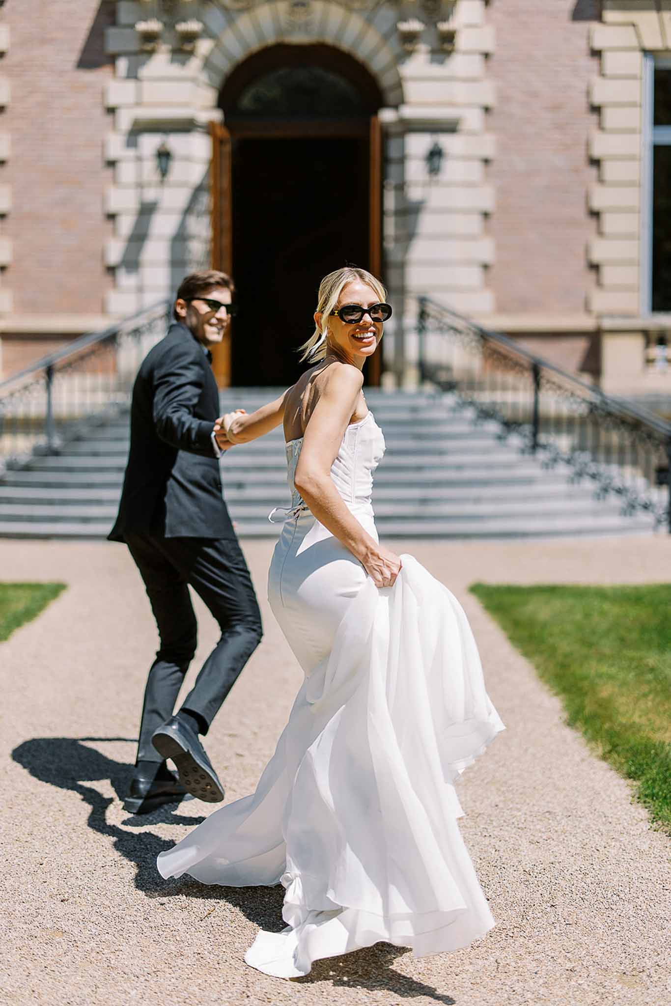 Laughing couple in sunglasses walking back toward camera on gravel path before manor entrance