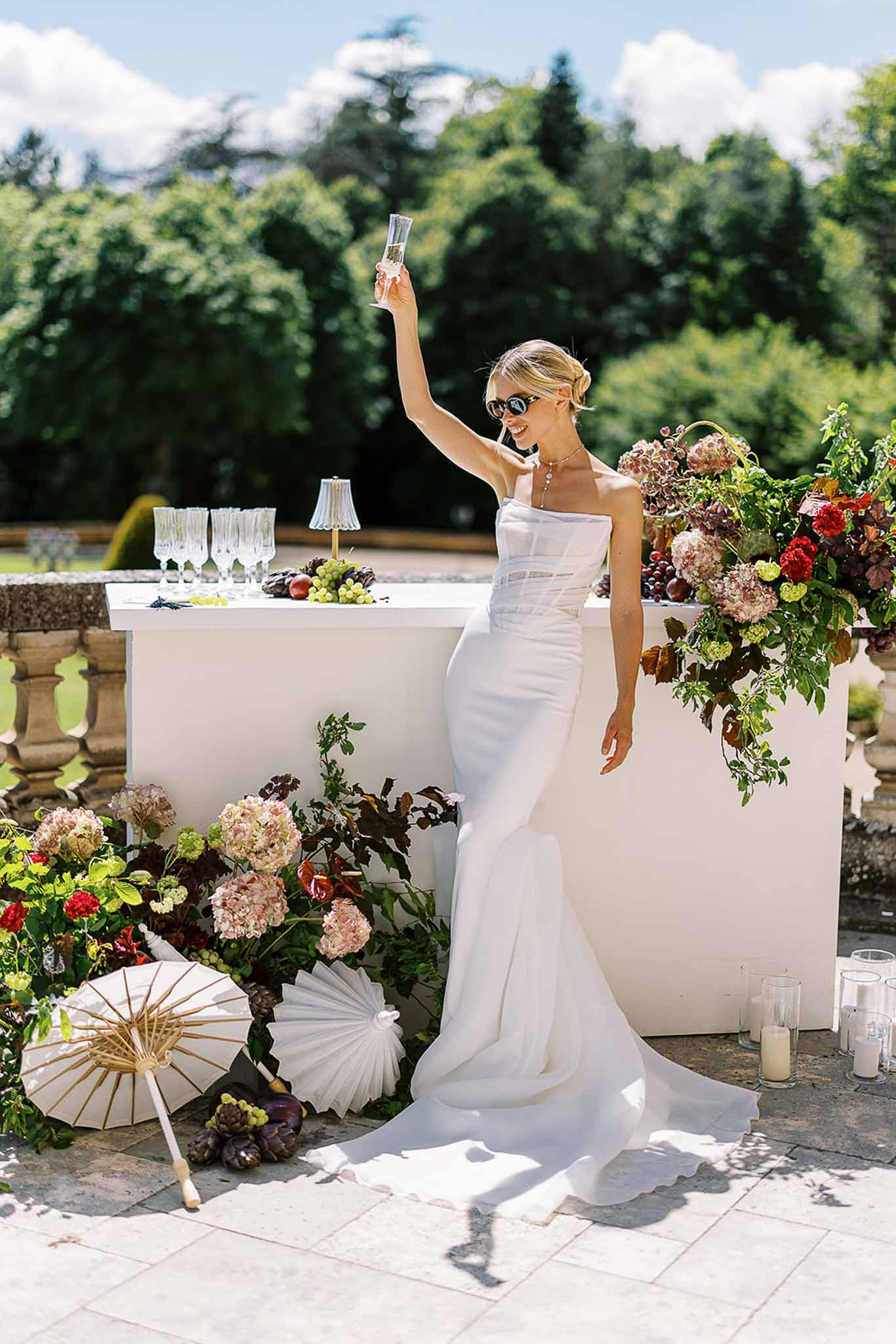 Bride raising champagne at white bar with mauve hydrangea and burgundy floral display on stone terrace