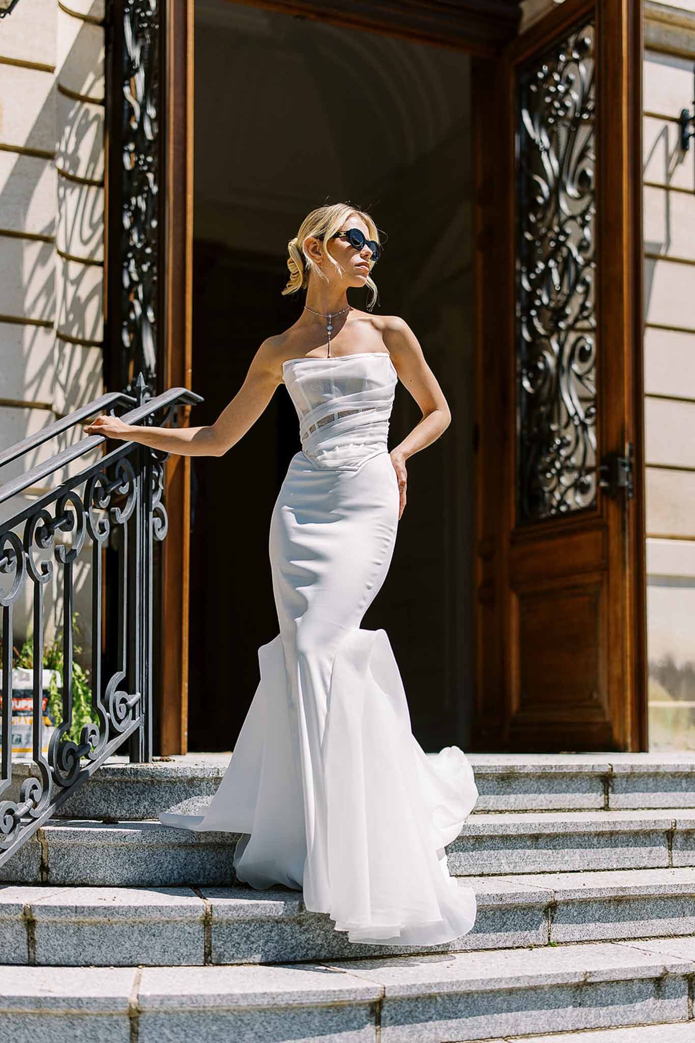 Bride in white strapless mermaid gown with sunglasses posing on stone steps in front of ornate wooden doors