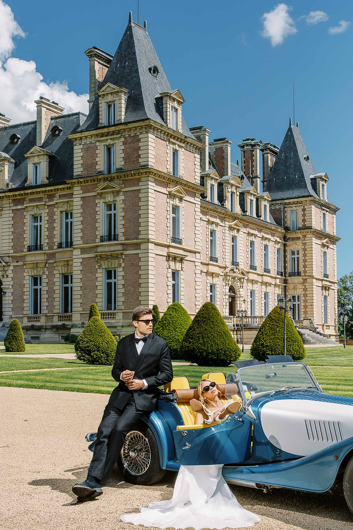 Couple posing with vintage blue roadster before grand chateau groom in tuxedo bride reclining in driver seat