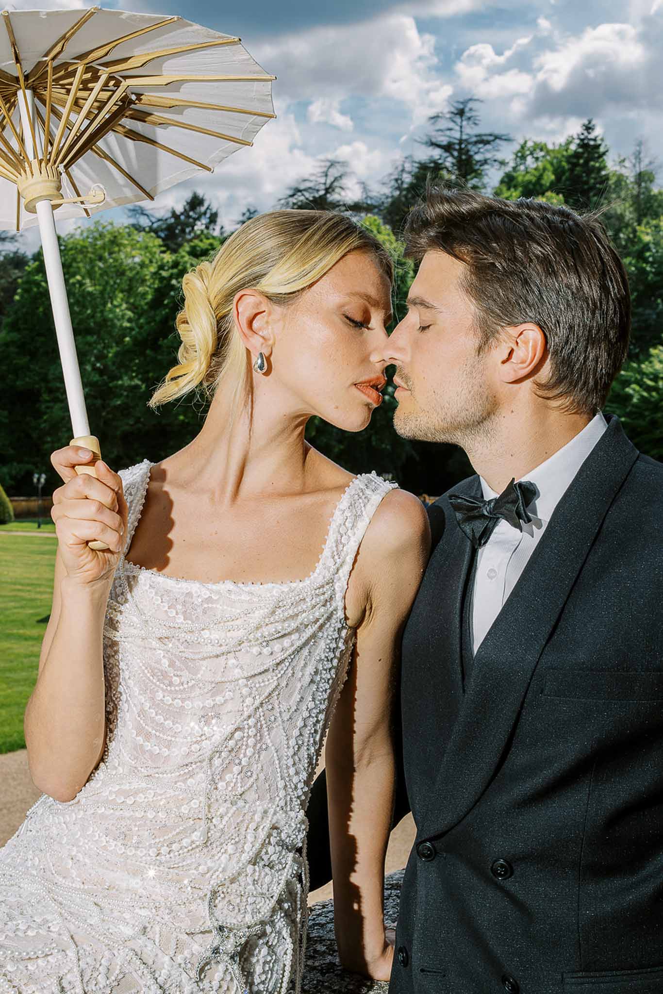 Close-up of bride in beaded ivory gown holding a white parasol near groom in charcoal suit in a garden