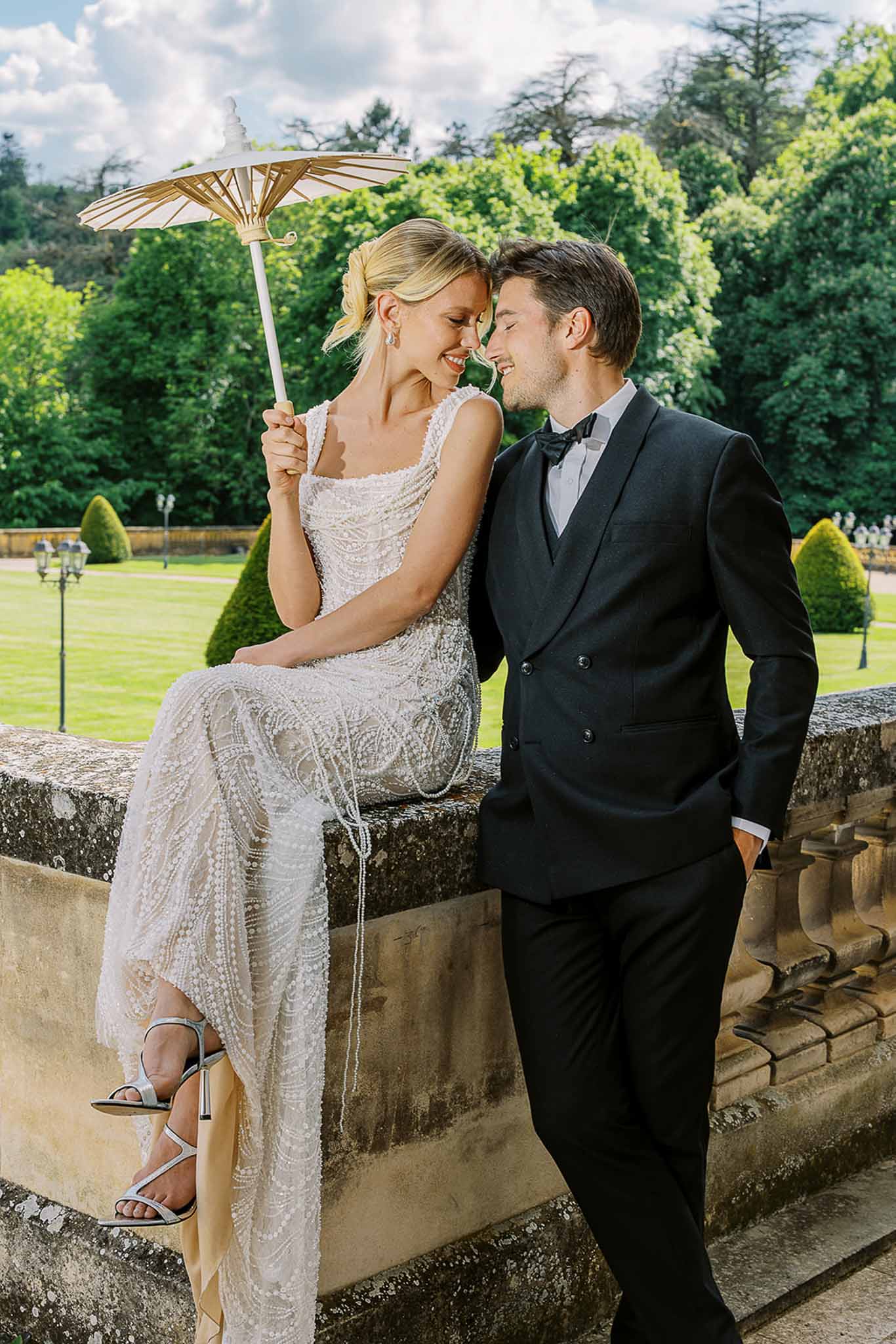 Bride in beaded ivory gown with parasol and groom in double-breasted tuxedo touching foreheads on stone balustrade