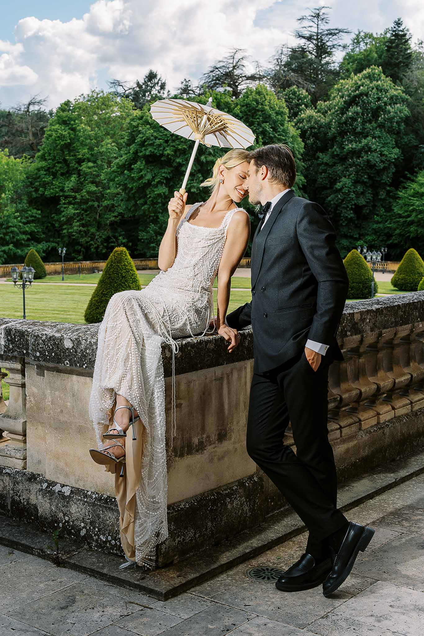 Bride in pearl-embellished gown seated on stone balustrade with groom in black tuxedo, formal garden behind