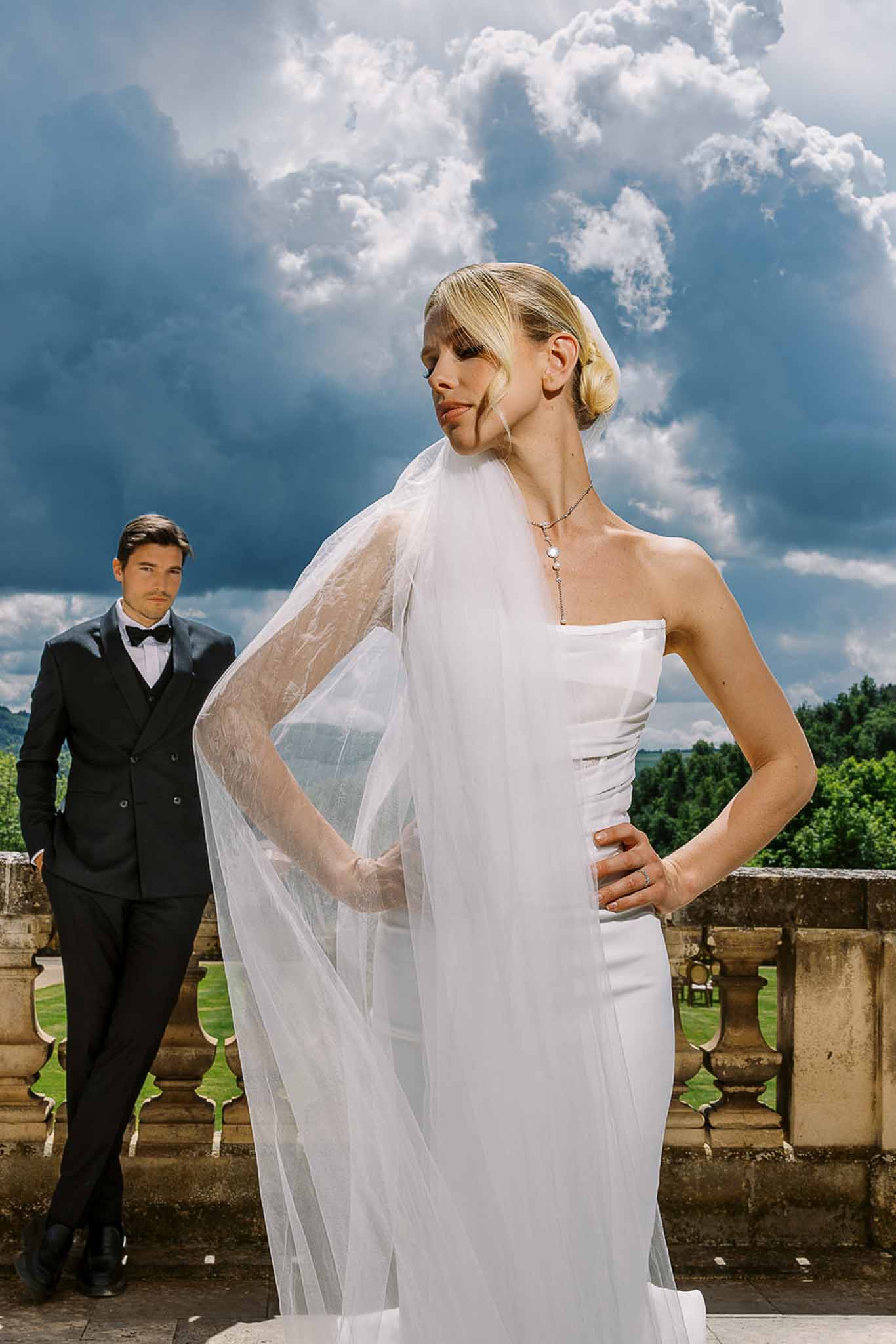 Bride in strapless gown and flowing veil poses on chateau terrace with groom in black tuxedo behind her