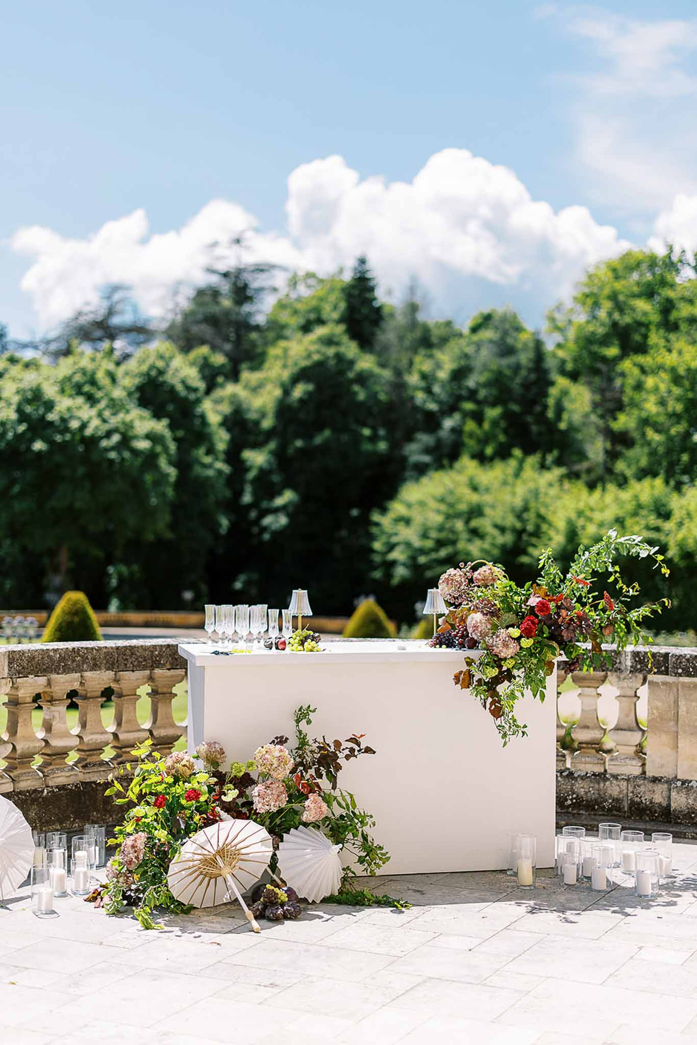 White bar counter with champagne glasses, dusty pink hydrangeas, burgundy florals, and paper parasols on terrace