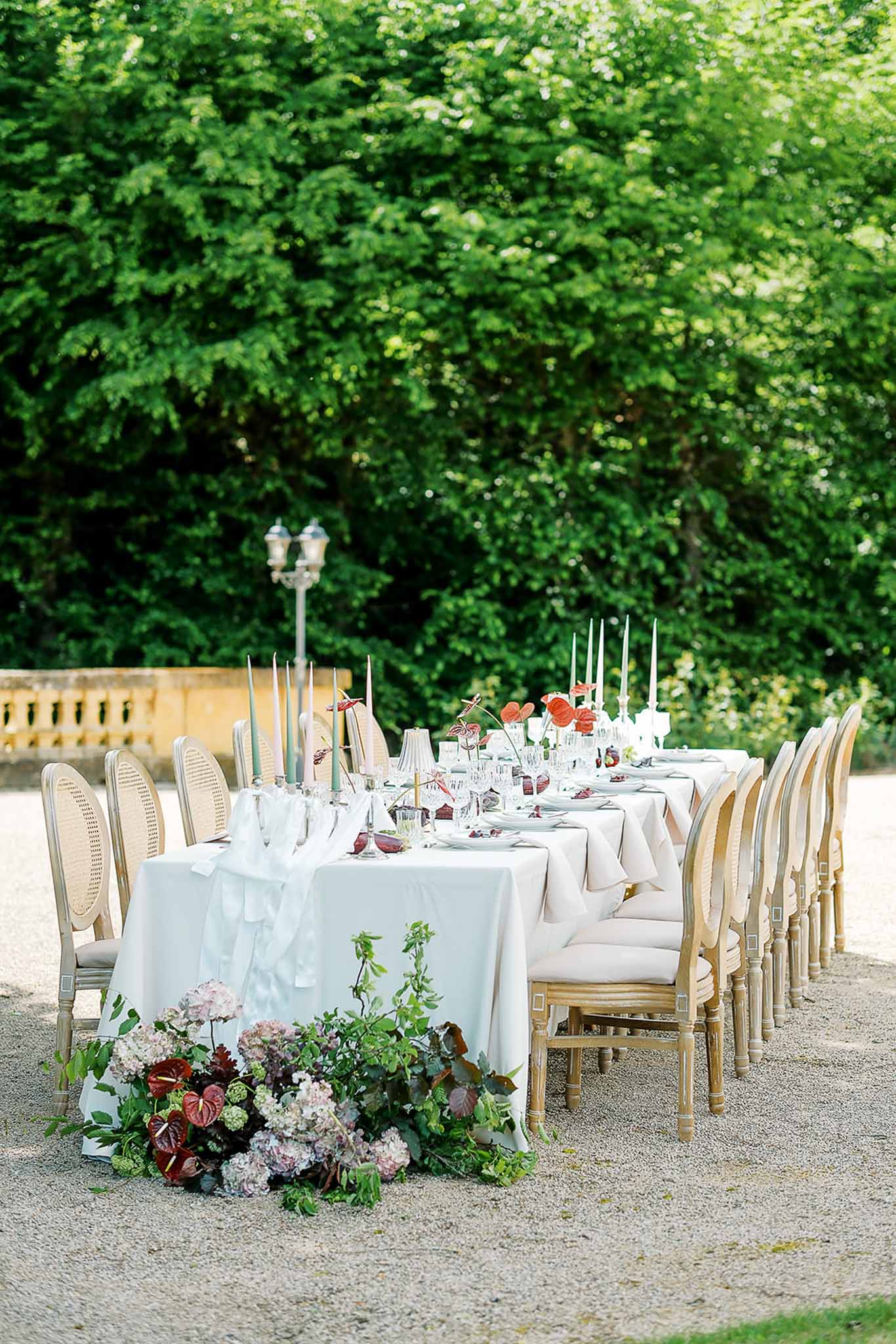 Long table with red anthurium accents, sage and gold tapers, and Louis XVI cane-back chairs on gravel