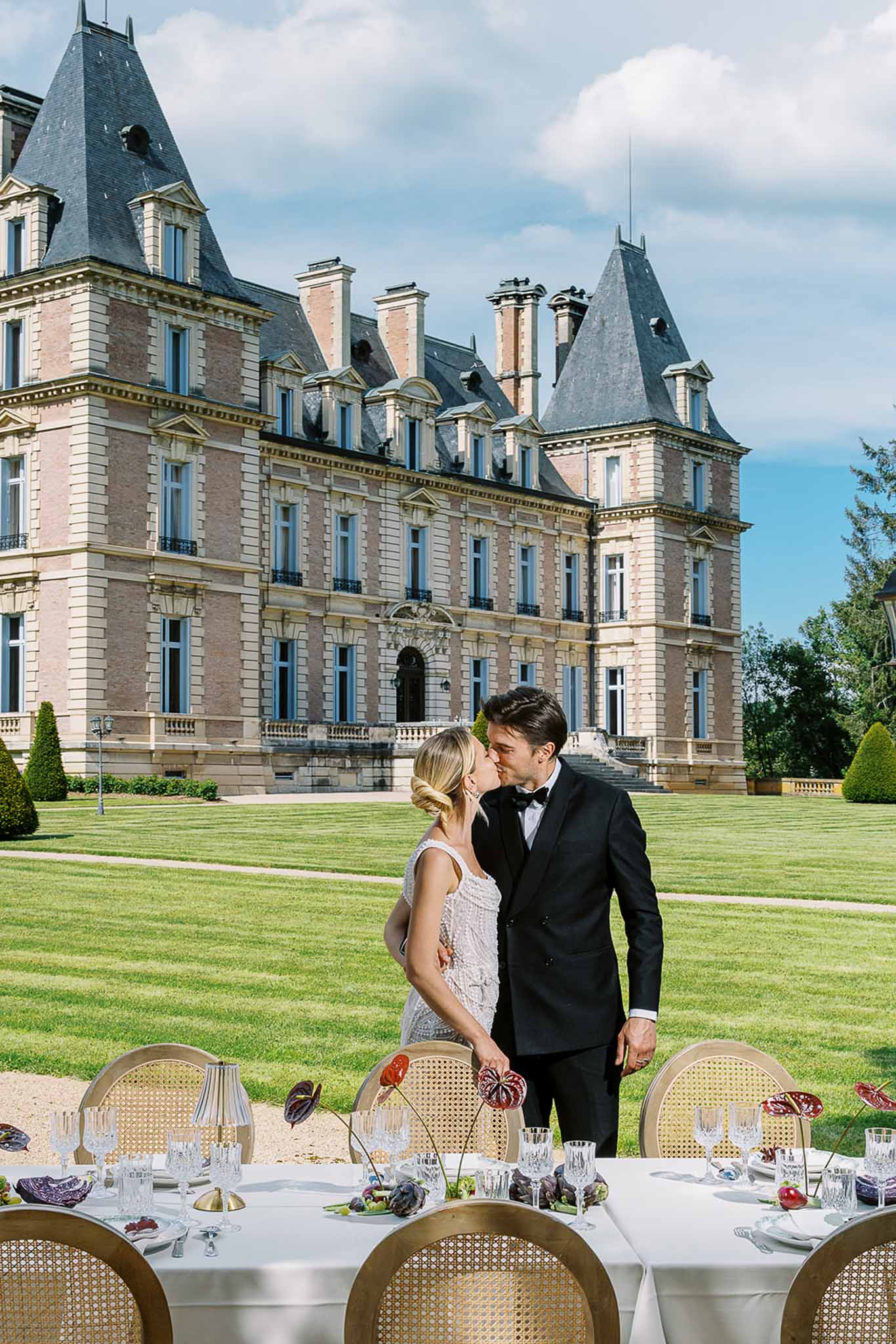 Bride and groom kissing behind styled reception table with gold candelabras and burgundy flowers in front of chateau