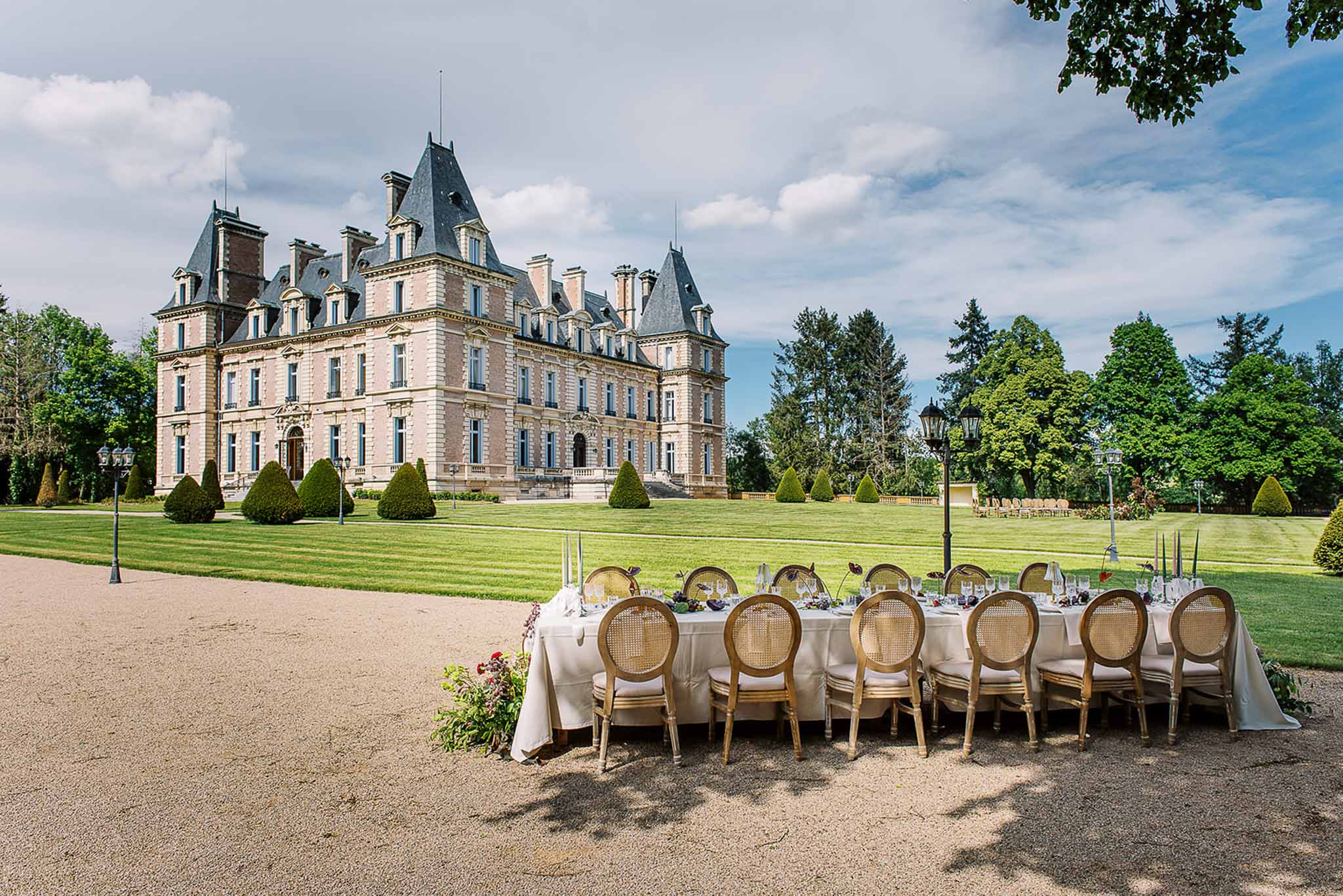 Long table with burgundy centrepieces and gold cane-back chairs on gravel drive before mansard chateau
