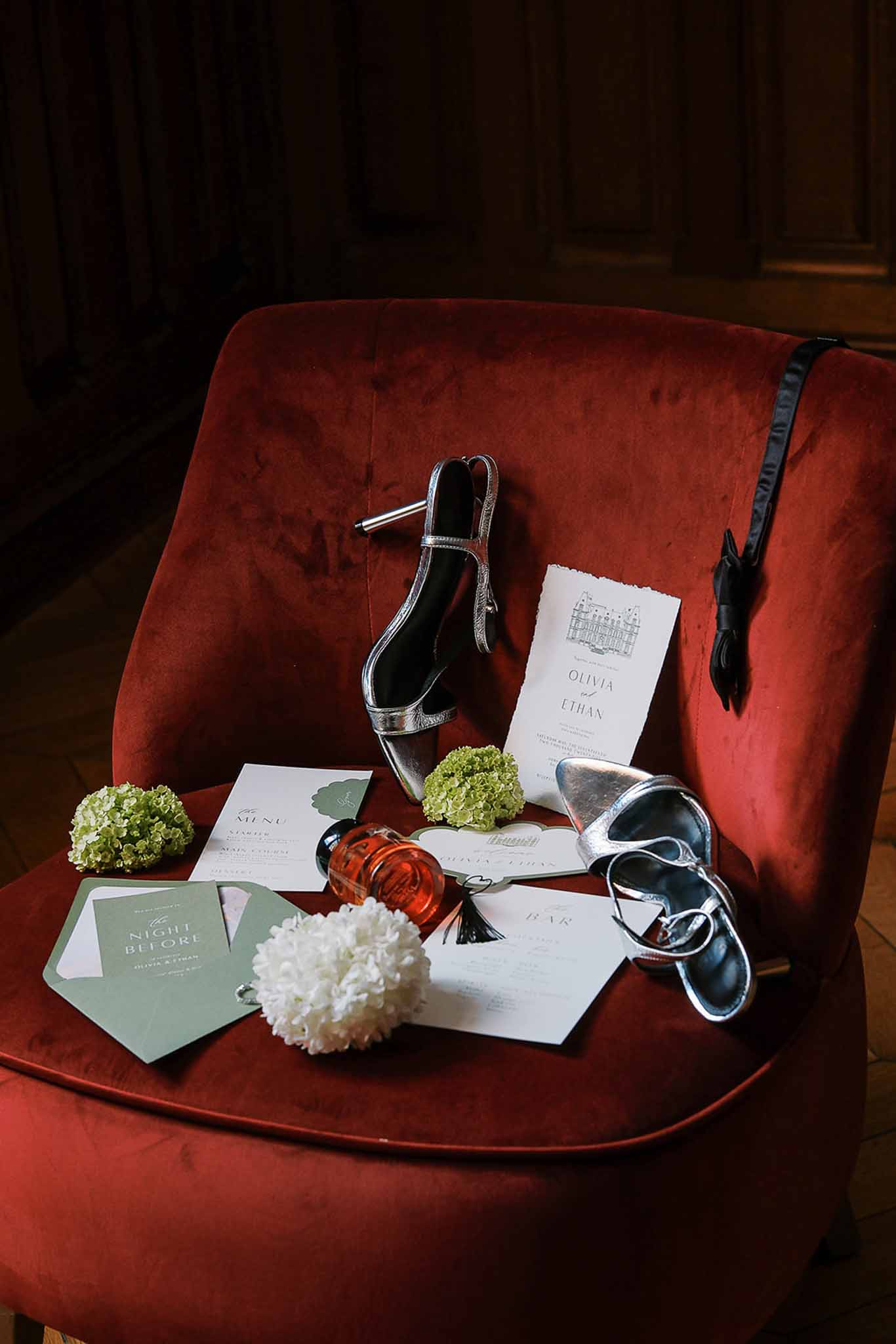 Wedding stationery flat lay with chateau illustration invitation, silver heels, and sage envelopes on red velvet chair