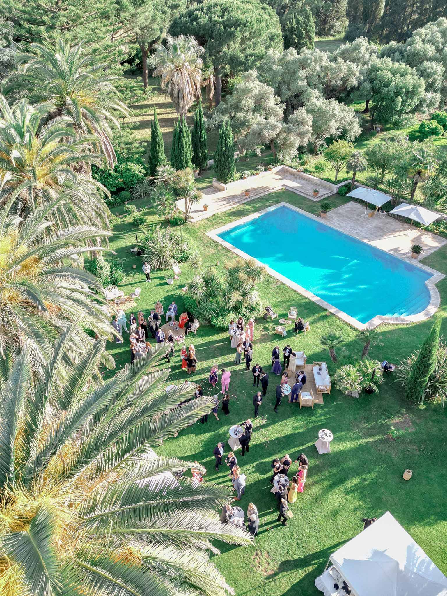 Aerial drone view of cocktail hour with 50 guests mingling around swimming pool and lawn at Mediterranean villa