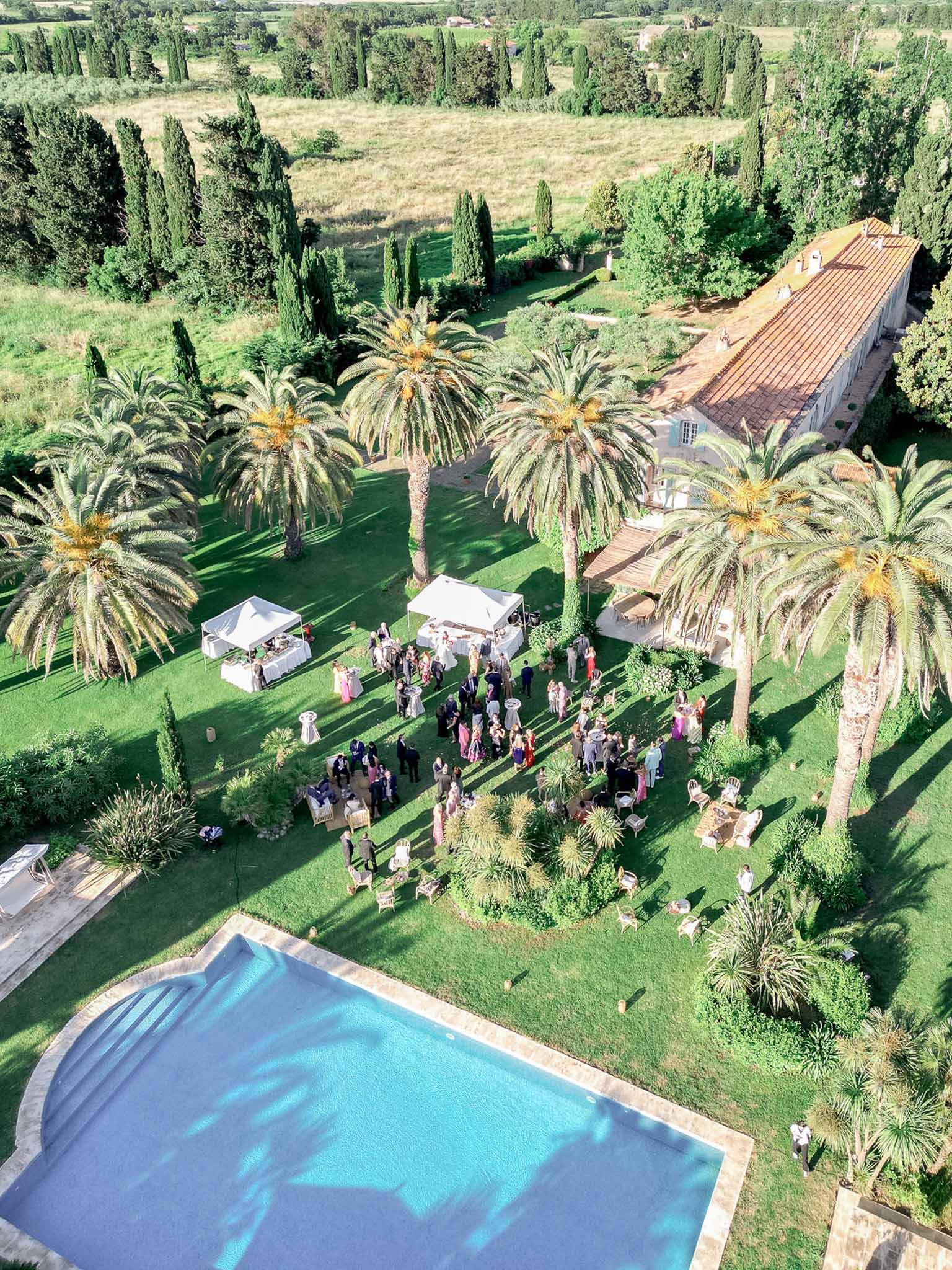 Aerial view of Provencal bastide cocktail hour with 70 guests, pool, palm trees, and white canopies