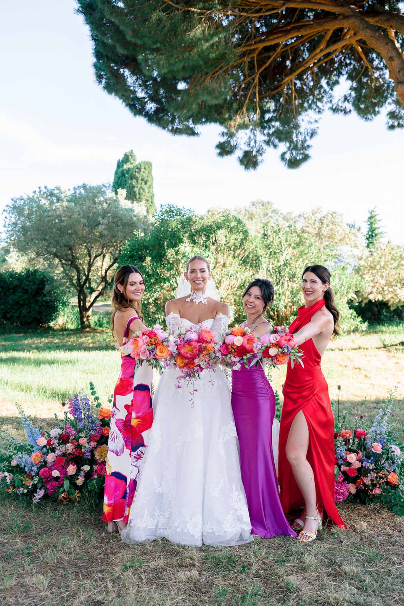 Bride with three bridesmaids in bold magenta, red, and floral gowns holding hot pink and coral rose bouquets