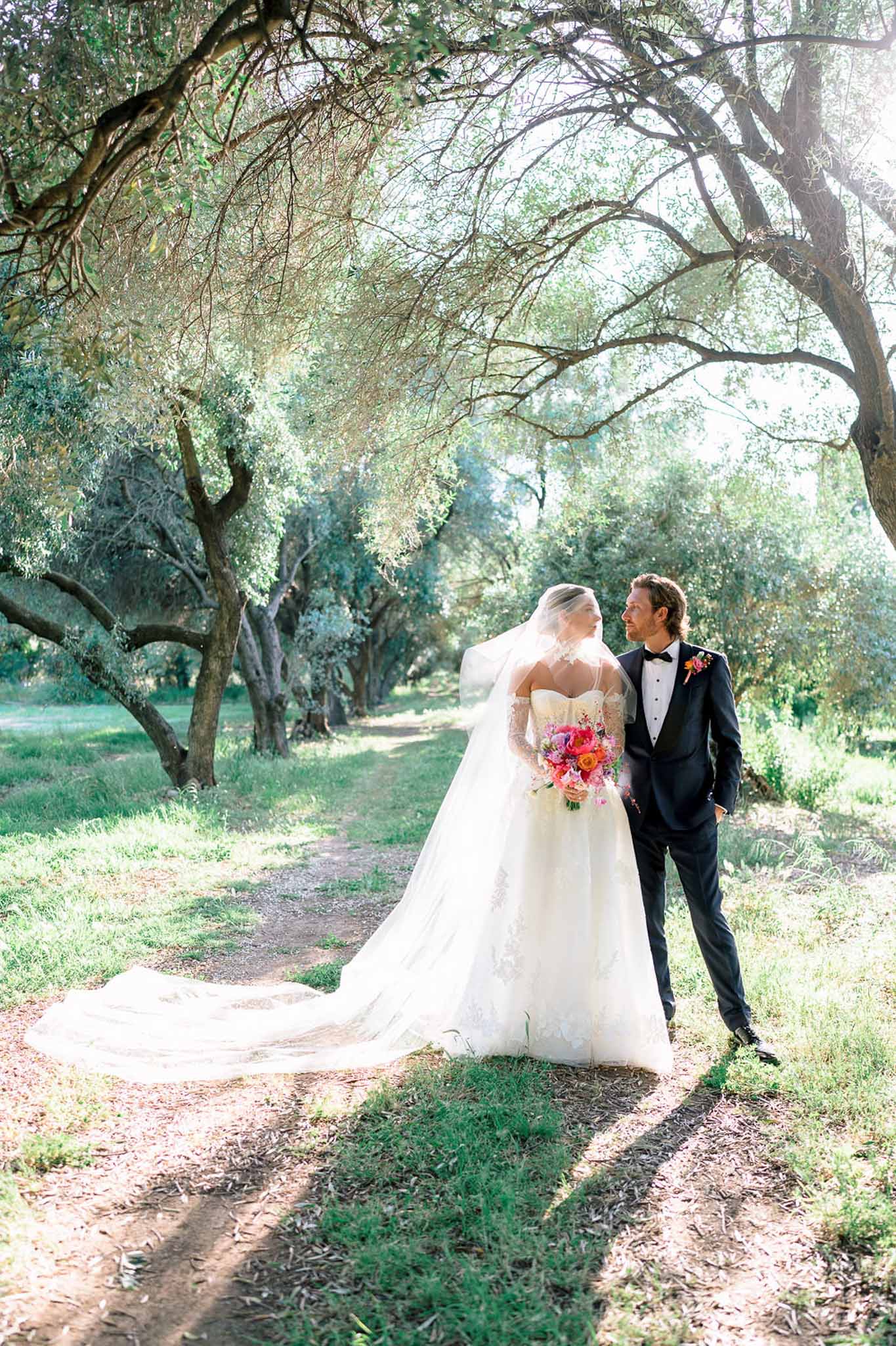 Bride in lace ballgown and groom in navy tuxedo on tree-lined olive grove path