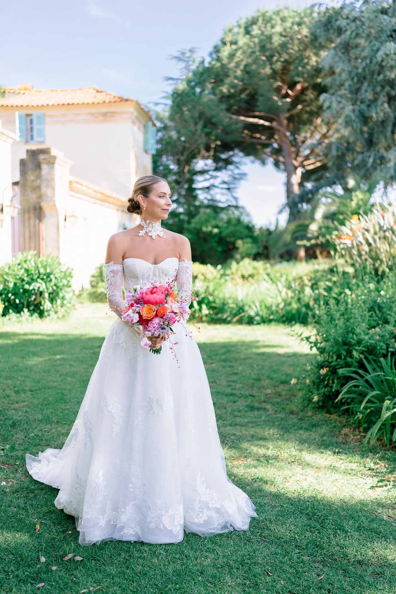Bride in off-the-shoulder lace ball gown with hot pink peony and orange rose bouquet on Provencal lawn