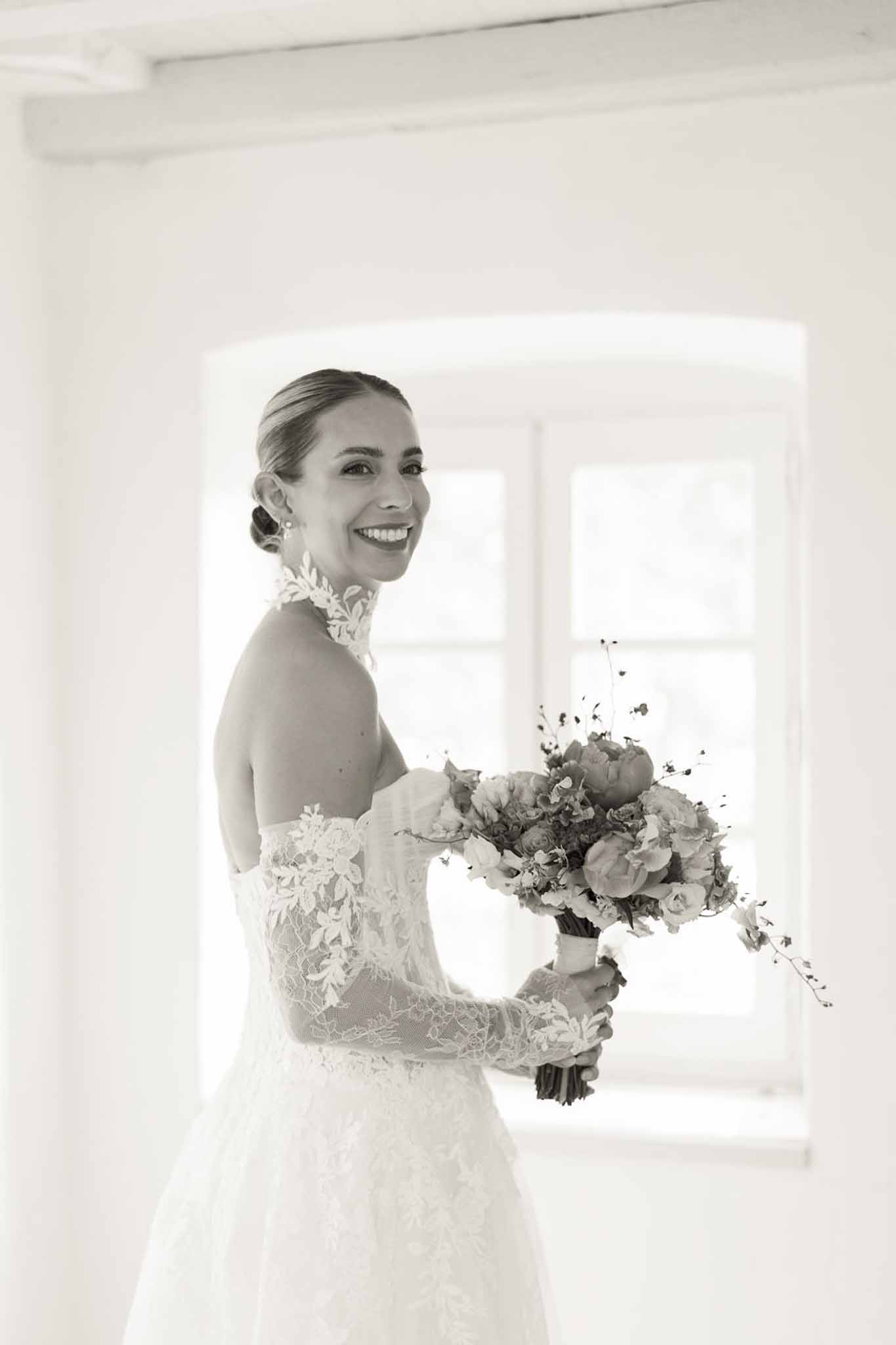 Black-and-white bridal portrait with bride in off-the-shoulder lace gown holding peony bouquet by arched window