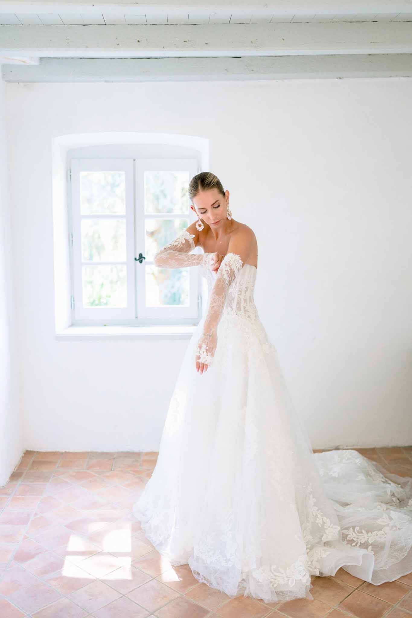Bride in off-shoulder ivory ball gown with lace sleeves and cathedral train standing in whitewashed Provencal room