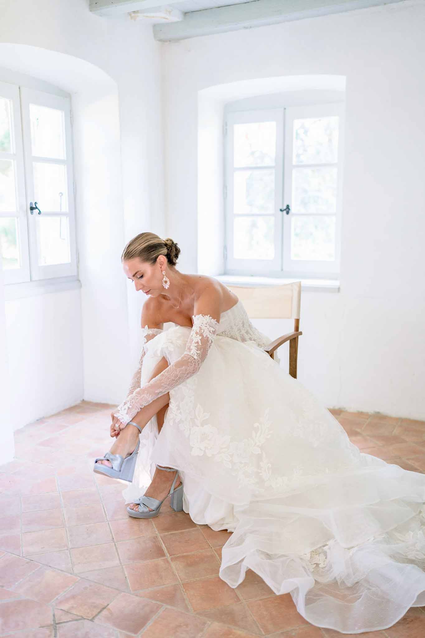 Bride in off-shoulder lace ballgown fastening powder-blue satin bow heels in Provencal whitewashed room