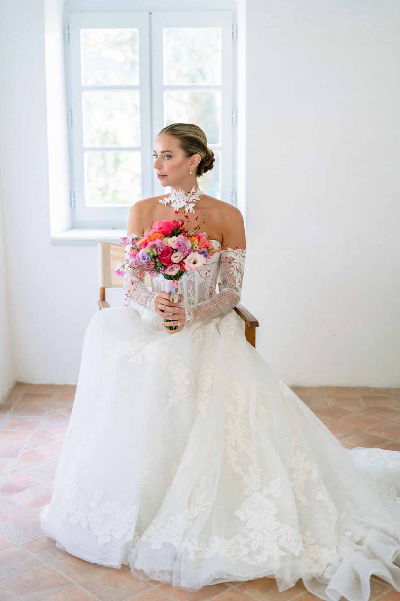 Bride seated in ivory lace ballgown with high collar holding hot pink peony coral rose and lavender bouquet by window