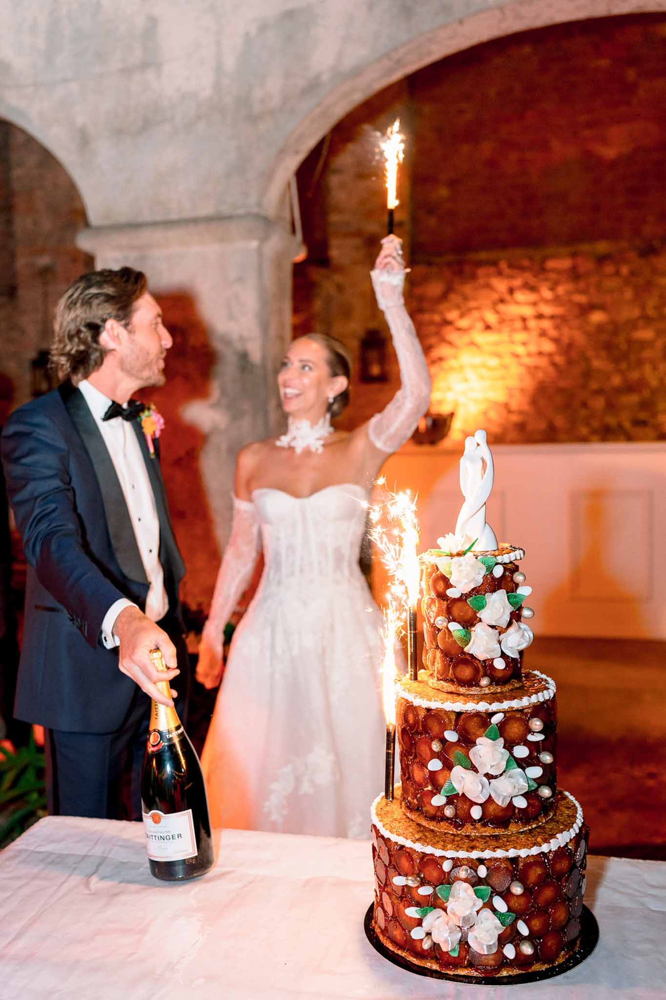 Bride with sparkler and groom with champagne beside croquembouche in stone-vaulted reception venue