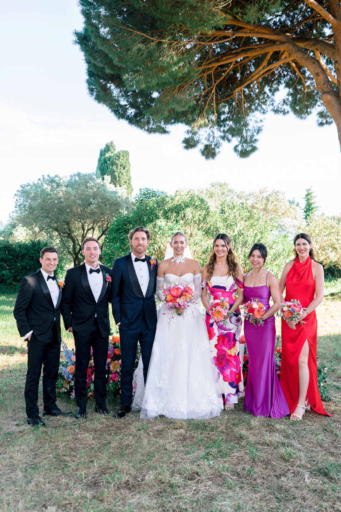 Bridal party of seven with hot pink and coral bouquets, mismatched colourful bridesmaids gowns, and black tuxedos