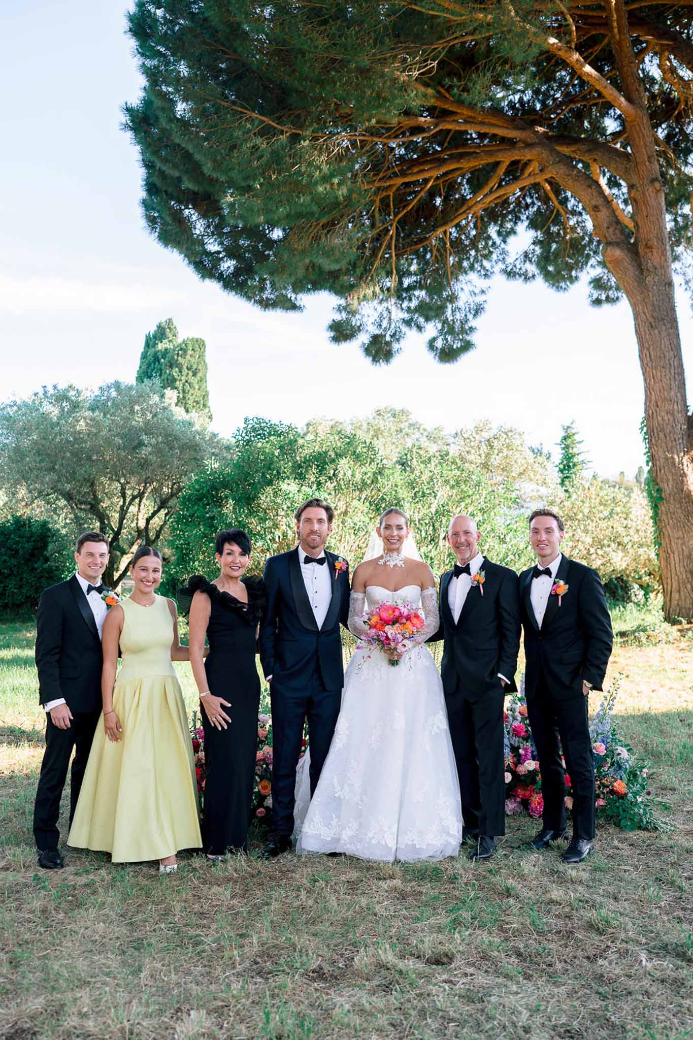 Seven-person group portrait with coral and orange bouquets, black tuxedos, yellow gown, and feathered black dress