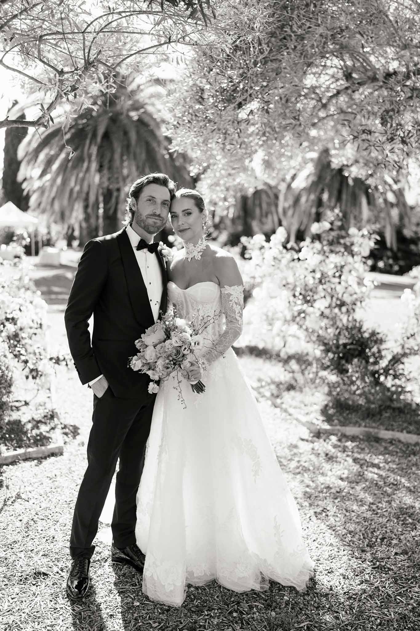Black and white couple in manicured garden bride in lace applique gown with trailing bouquet groom in tuxedo