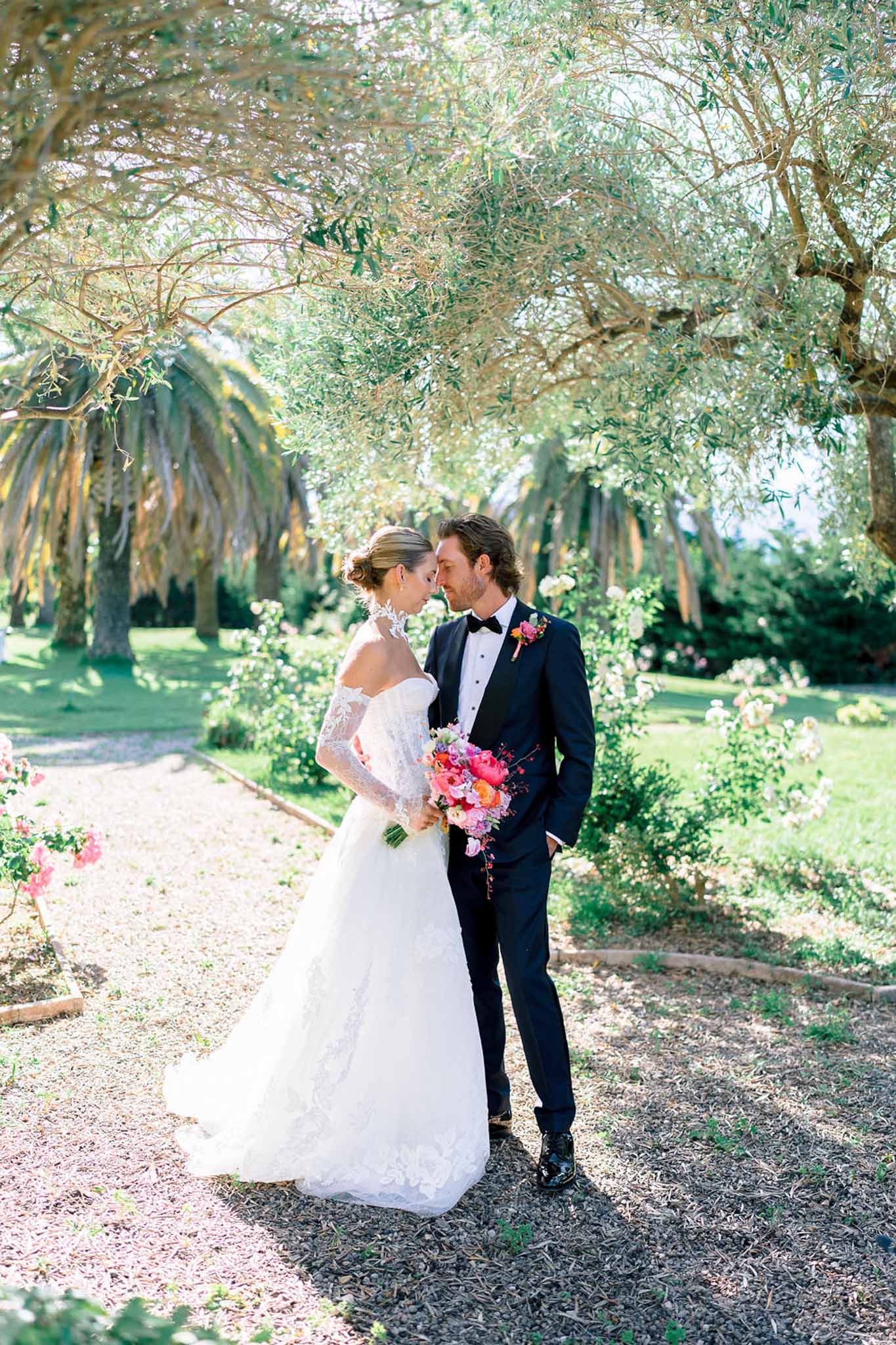 Bride in white lace ball gown and groom in navy tuxedo touch foreheads holding a hot pink and coral bouquet in a garden