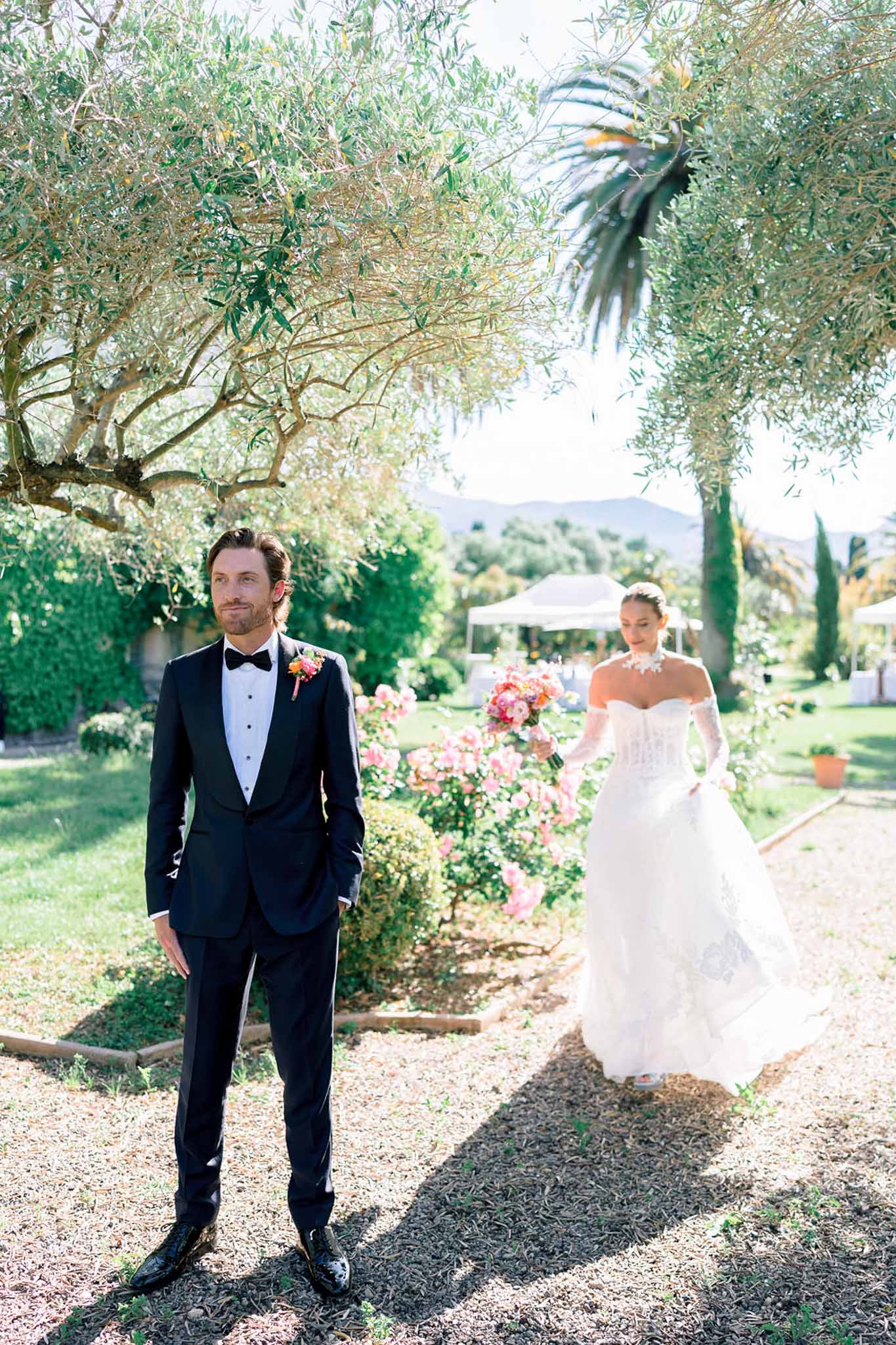 Bride in off-shoulder lace gown approaches groom in navy tuxedo along a garden path during first look at a Provencal estate