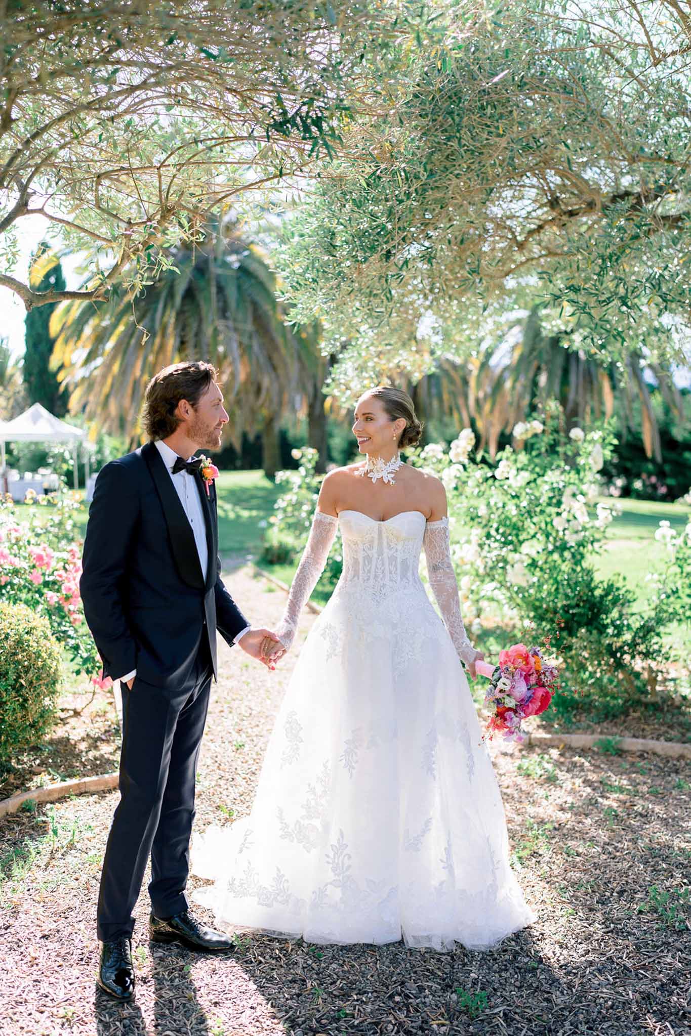 Bride in white lace off-the-shoulder gown and groom in black tuxedo holding hands in a garden with tree canopy above