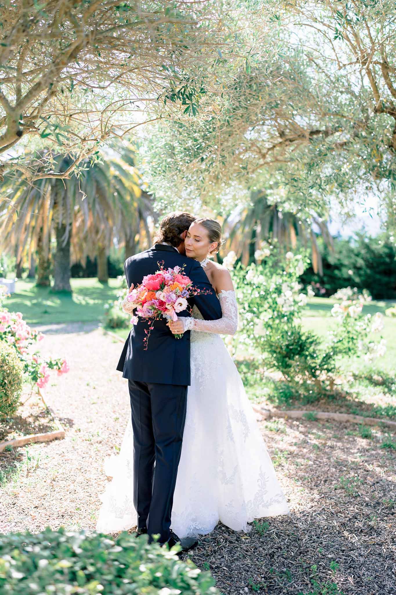 Bride looking over groom's shoulder holding hot pink peony and coral ranunculus bouquet in olive grove