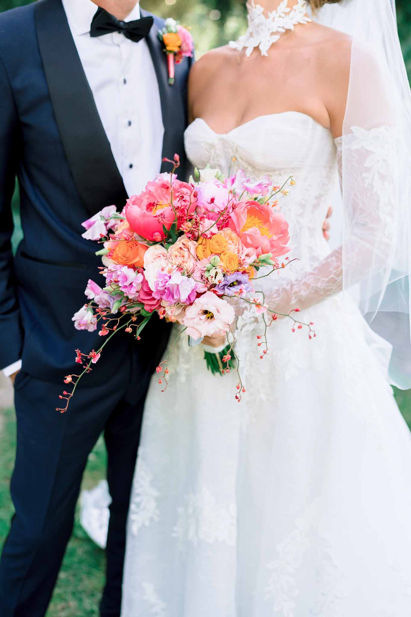 Close-up of bride and groom torsos with wild garden bouquet of hot pink peonies and coral roses