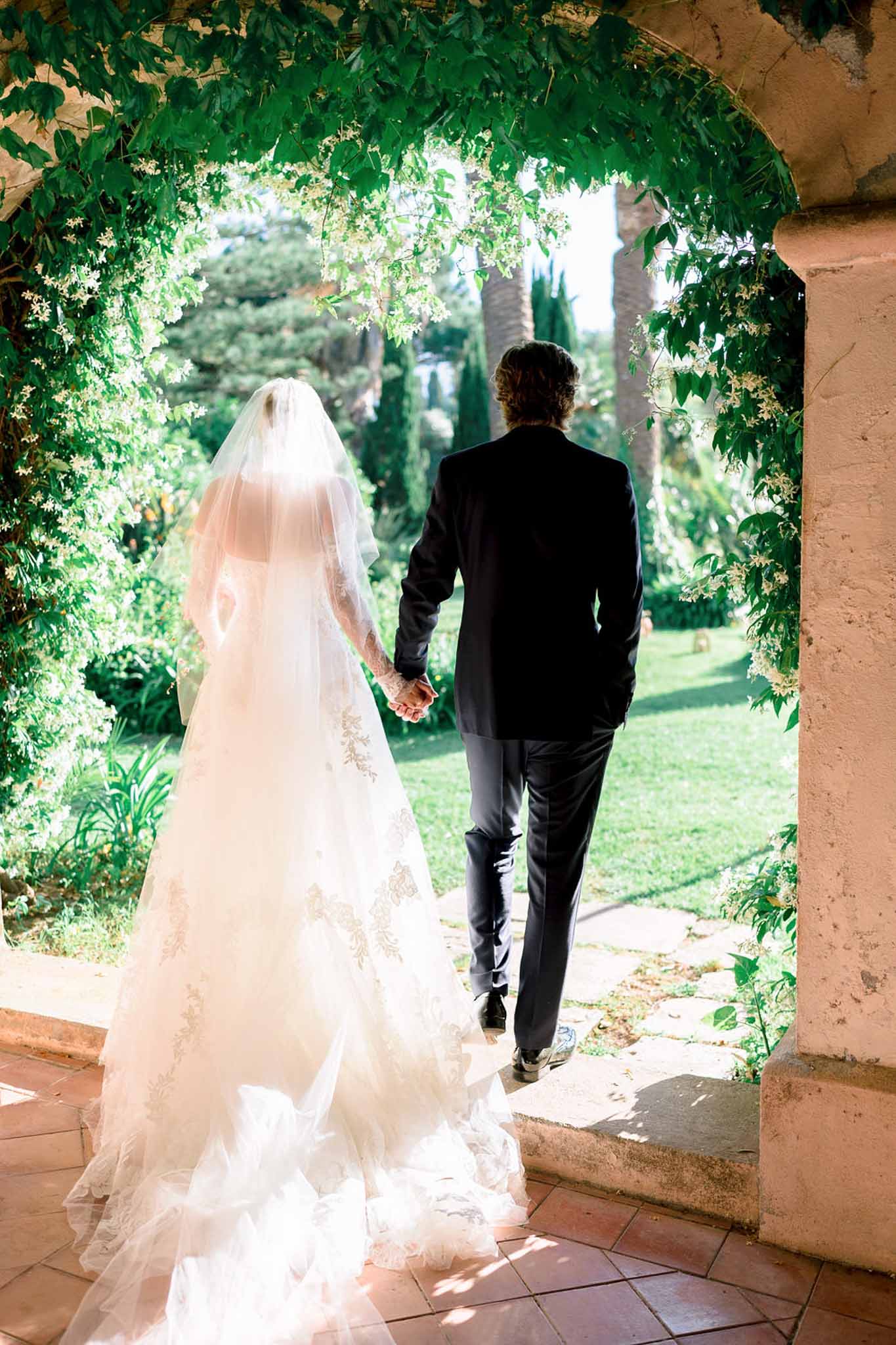 Bride and groom walking hand-in-hand through a vine-covered stone archway toward a sunlit formal garden