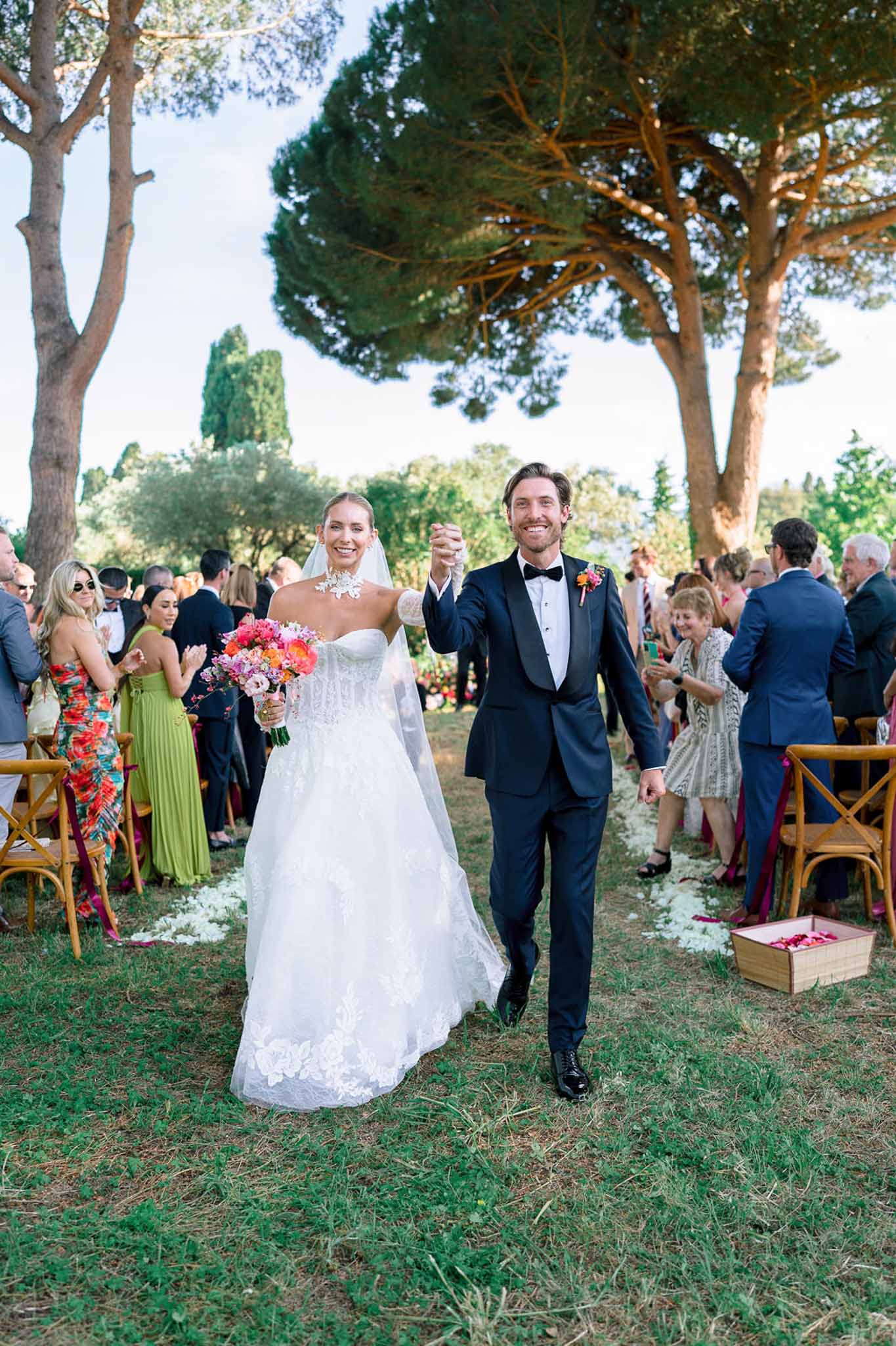 Couple raising hands after ceremony with hot pink and coral bouquet, white petal-scattered garden aisle