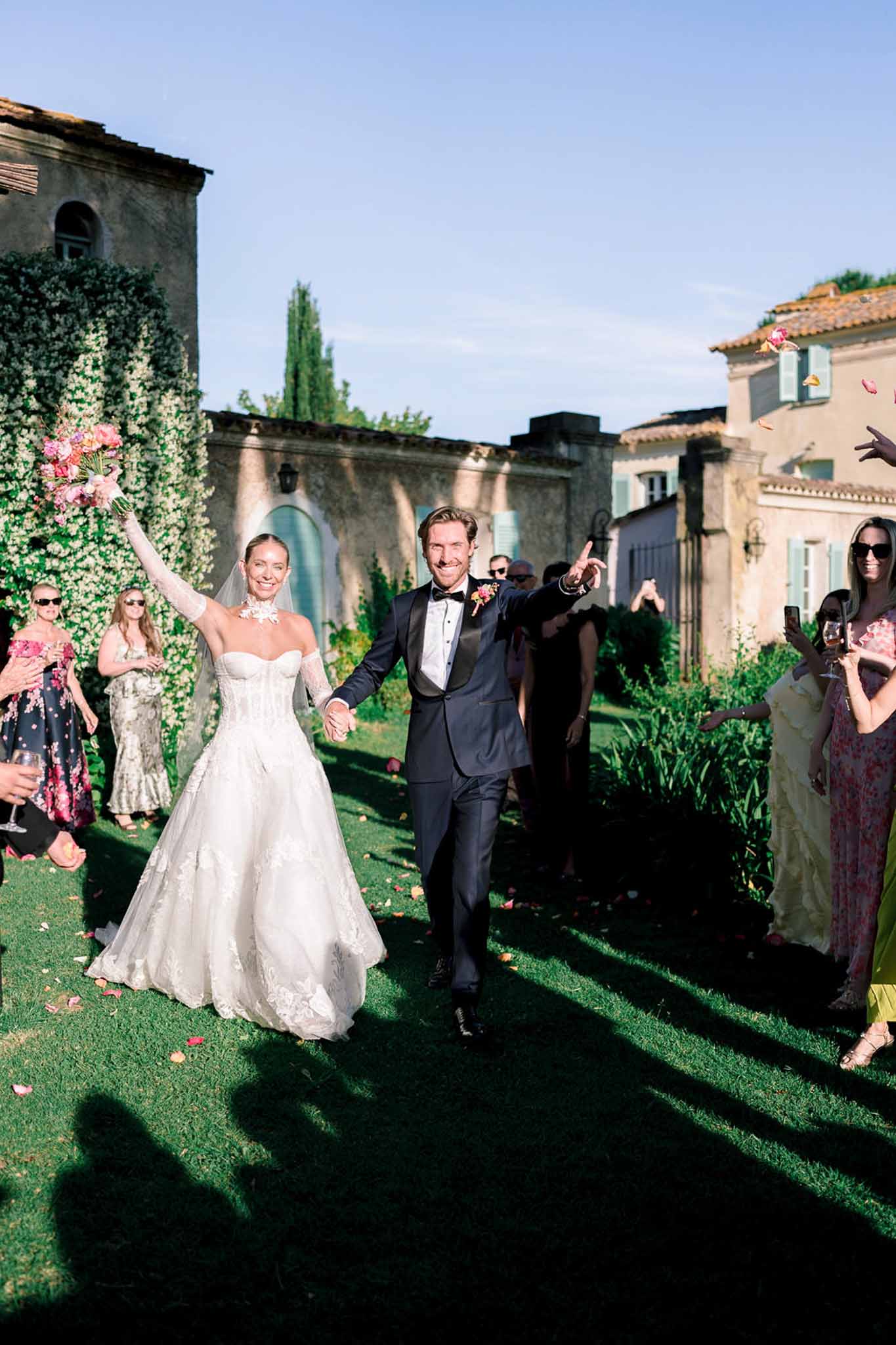 Couple's celebratory exit with petal toss past Provencal buildings in golden afternoon light