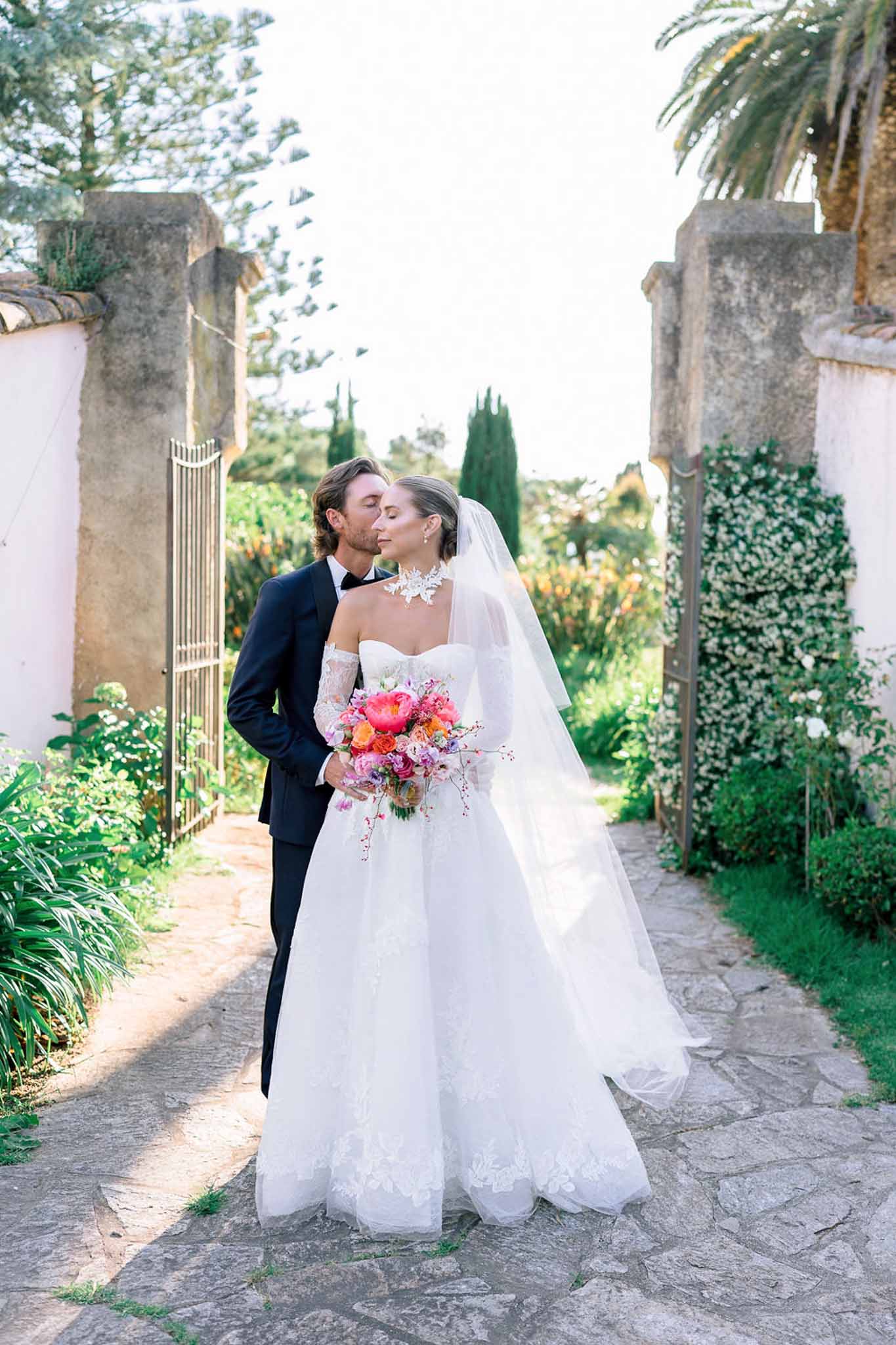 Groom kisses bride on cheek in walled garden as she holds colorful peony and rose bouquet