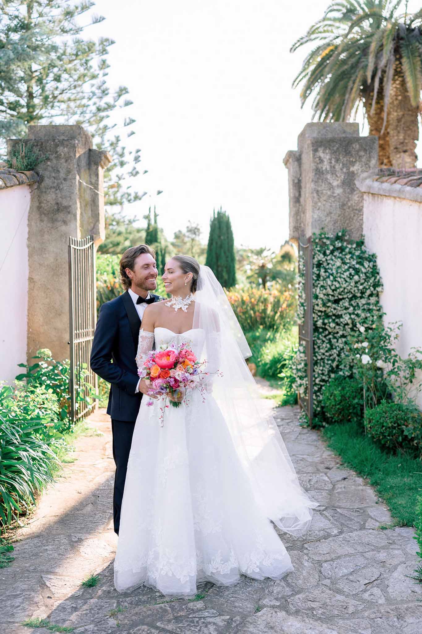 Couple at stone gate with bride holding hot pink peony and coral ranunculus bouquet and lace veil