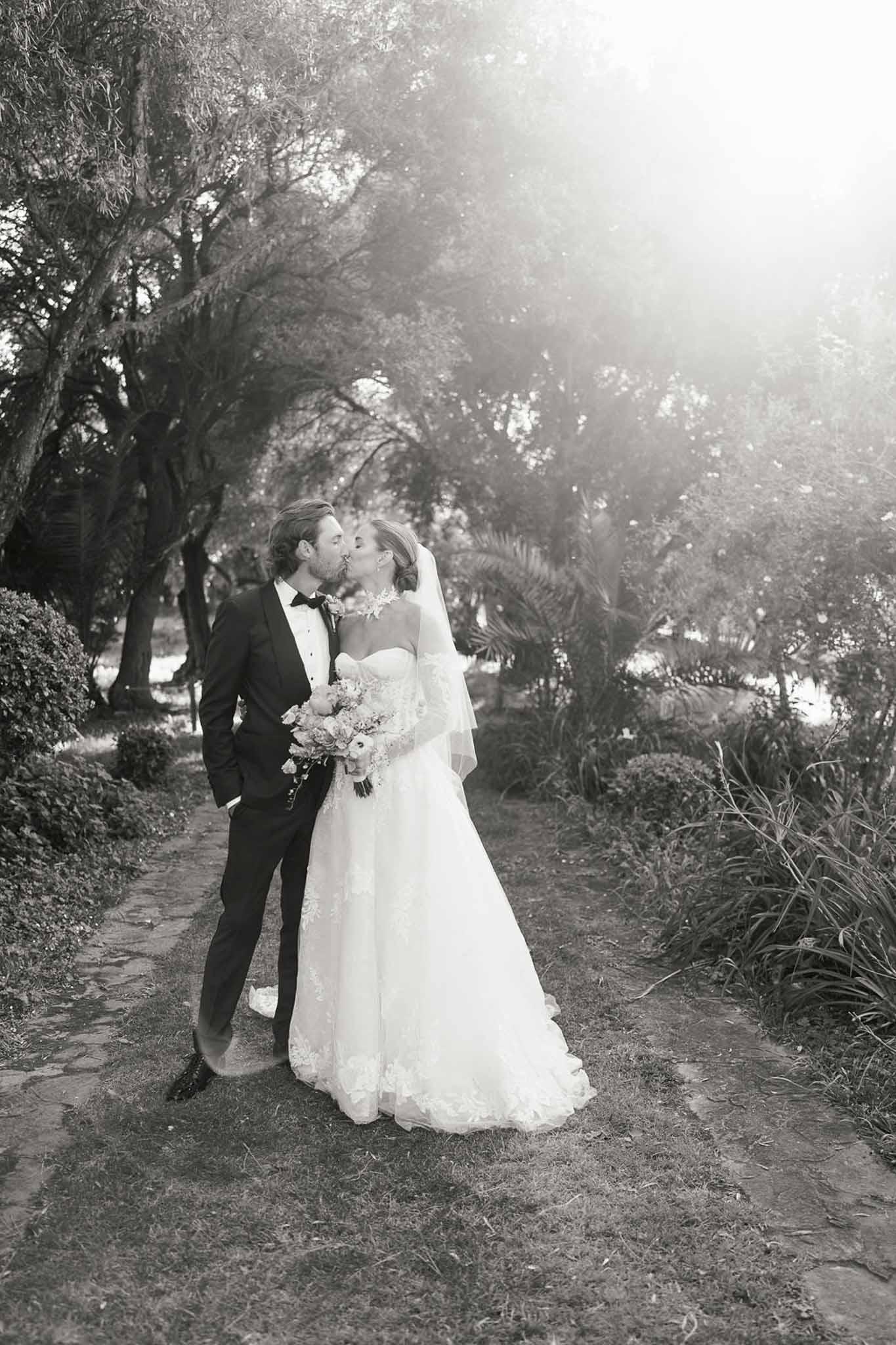 Black and white couple kissing on stone garden path bride in lace ball gown with elbow veil groom in tuxedo backlit