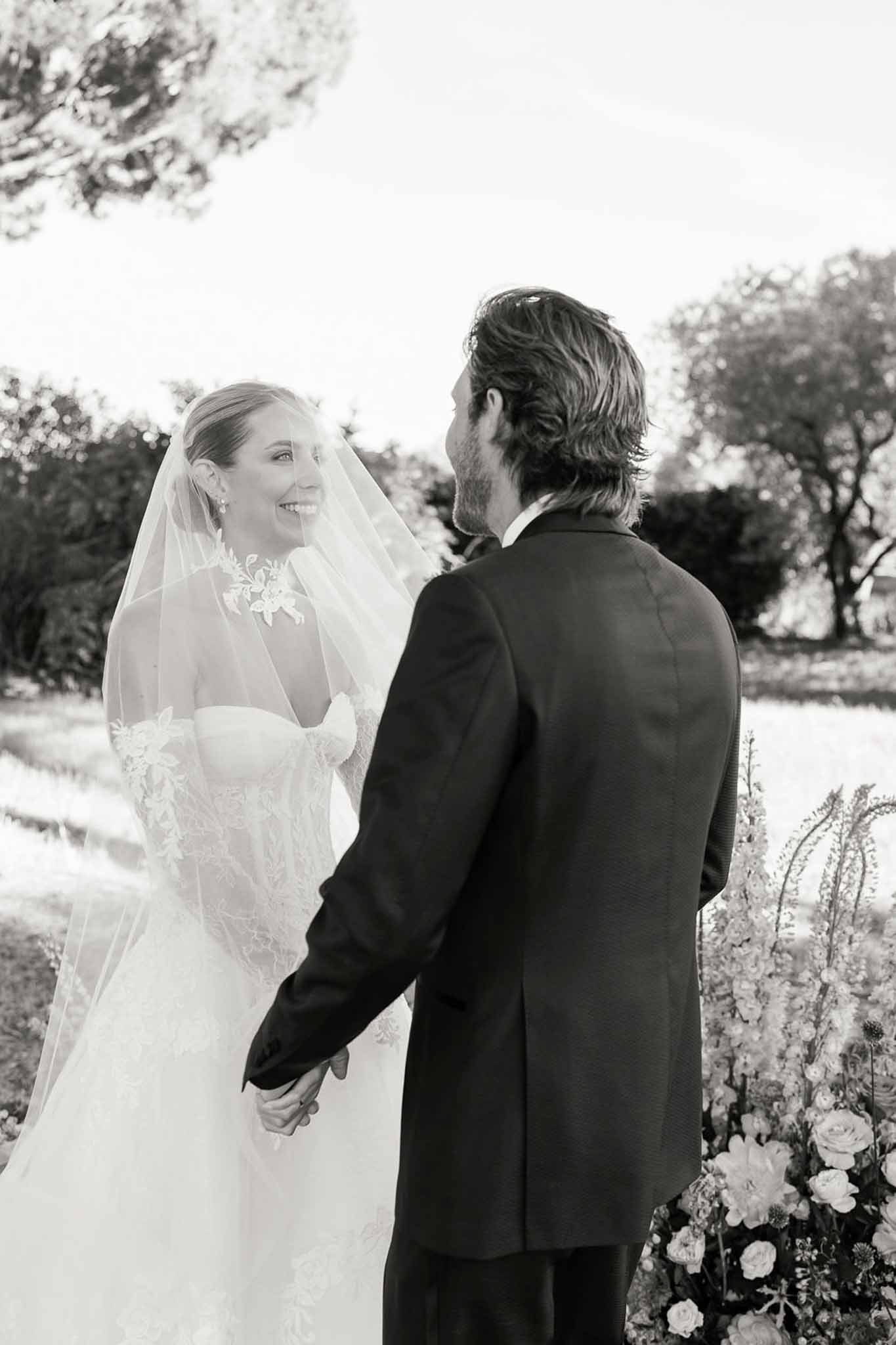 Black and white of bride smiling at groom during ceremony in strapless lace gown with floral cathedral veil
