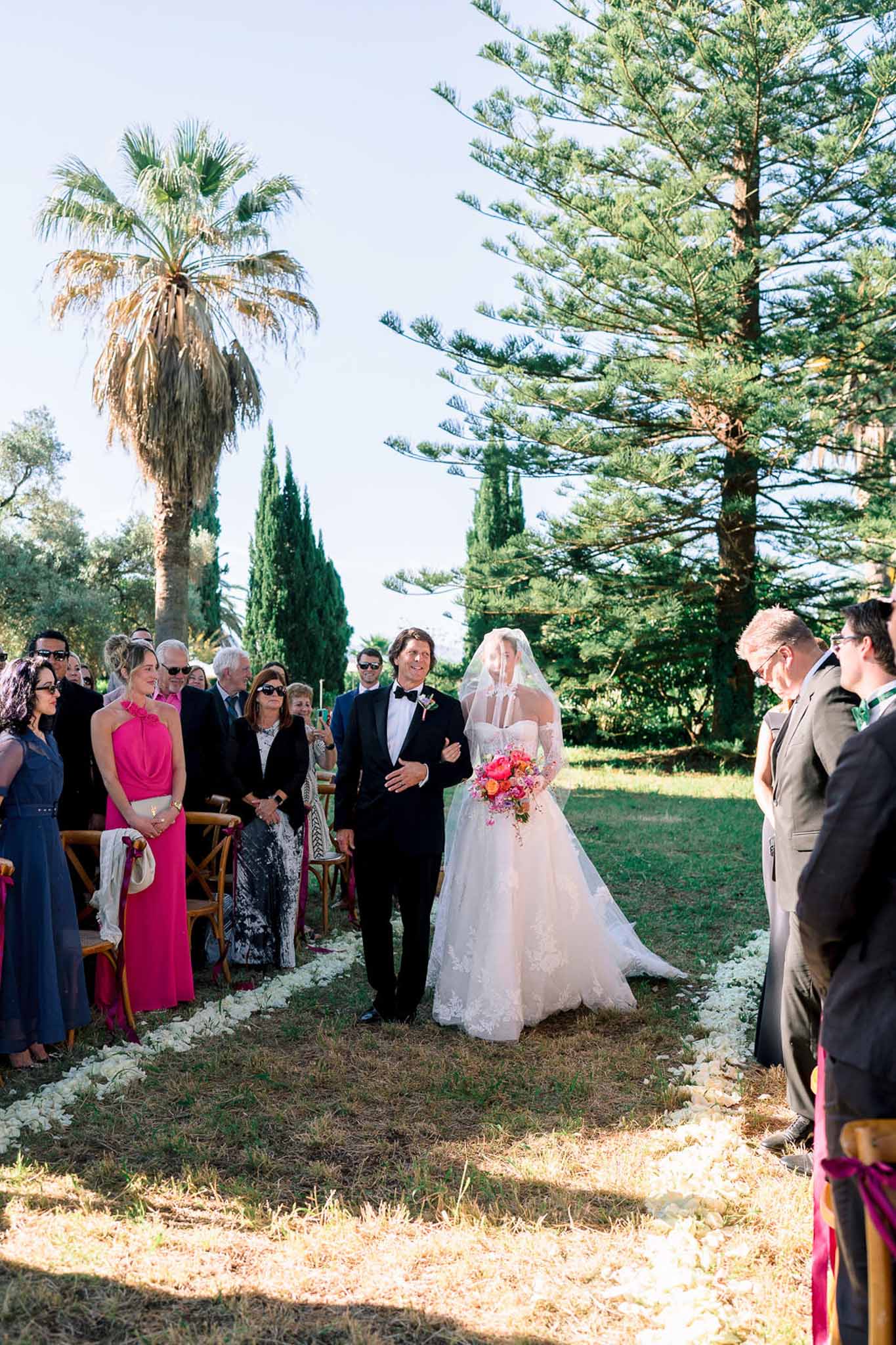 Bride walking aisle with hot pink bouquet past fuchsia-ribboned chairs in Mediterranean garden setting