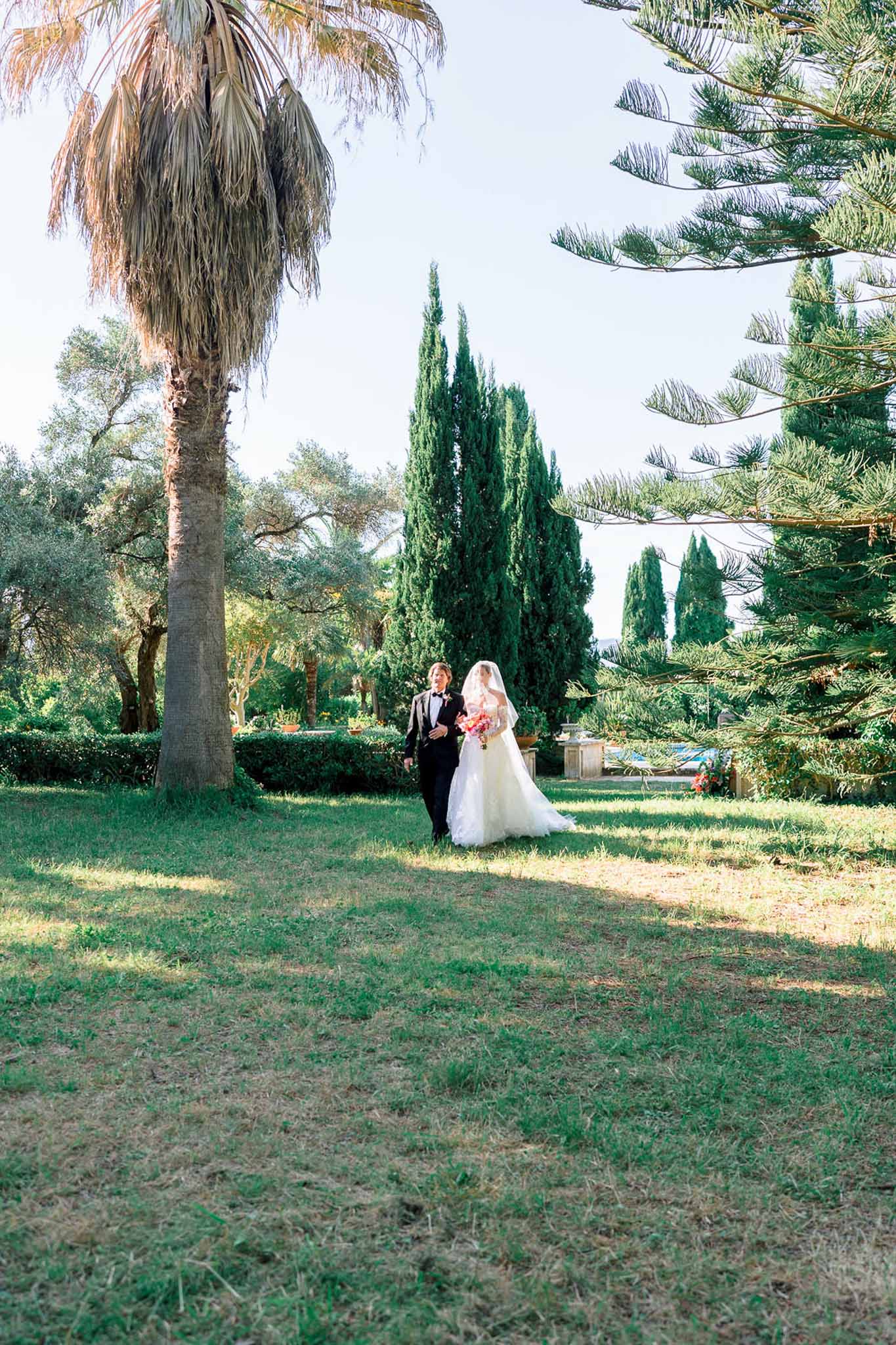 Bride with coral bouquet and groom in tuxedo walking through Mediterranean garden with cypress trees