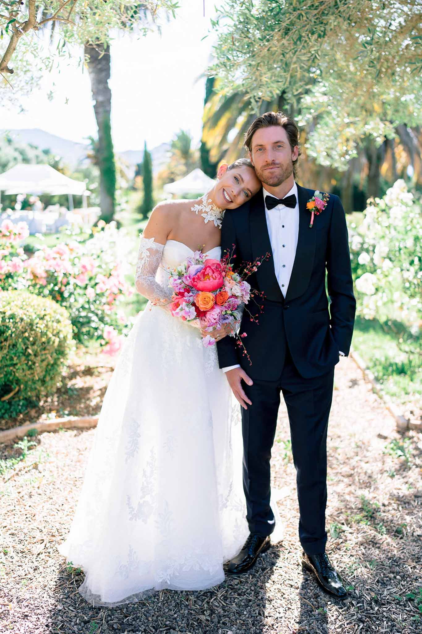 Bride leaning on groom's shoulder in garden holding vibrant pink peony and orange rose bouquet