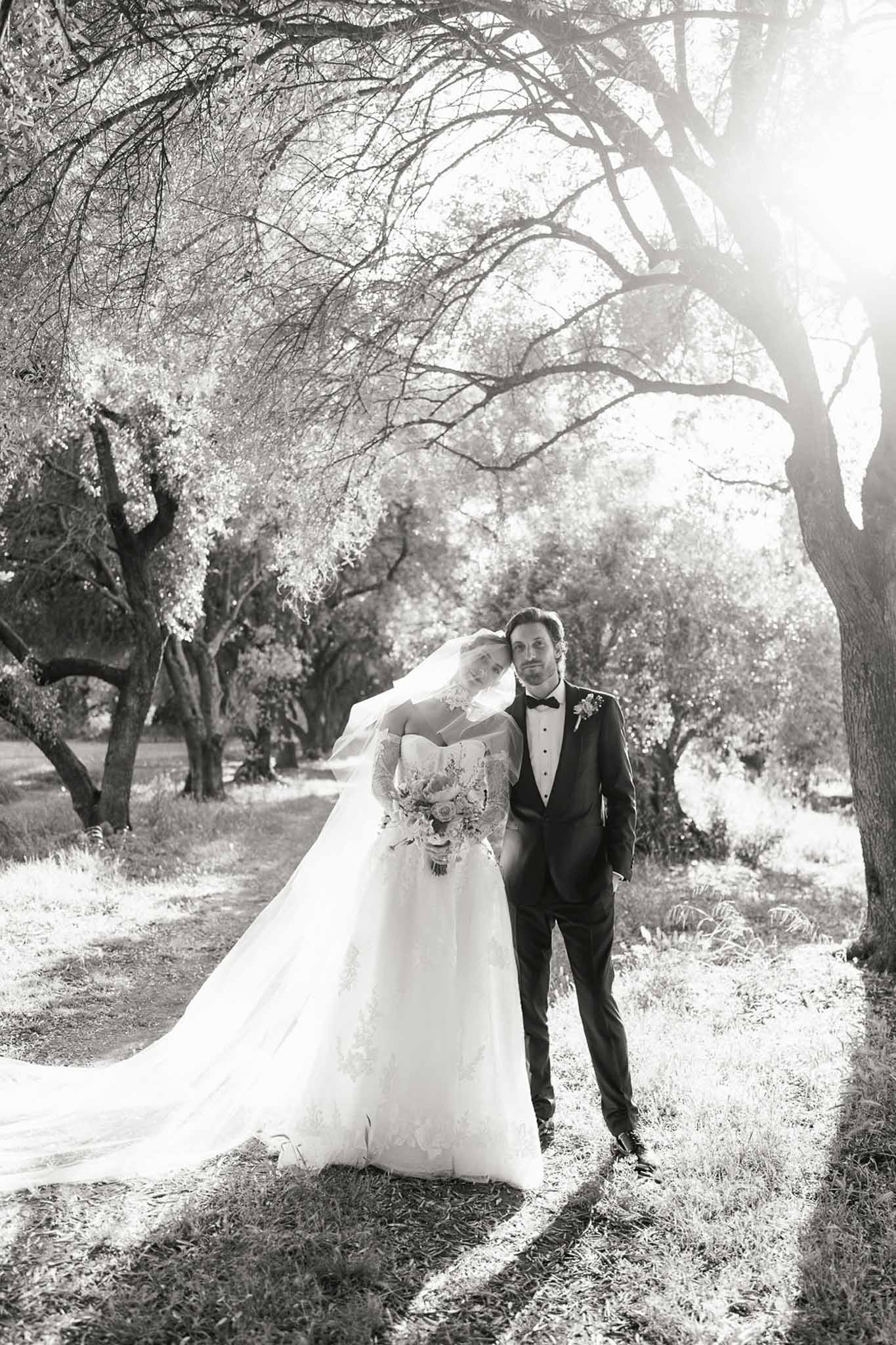 Black and white portrait of bride and groom walking along tree-lined path at French estate with backlit veil