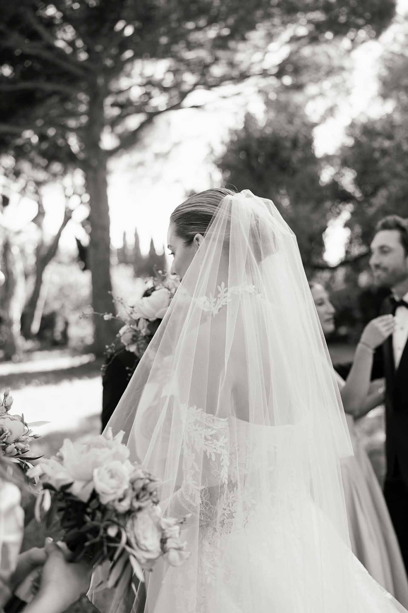 Black-and-white portrait of bride from behind showing lace-trimmed cathedral veil and garden rose bouquet