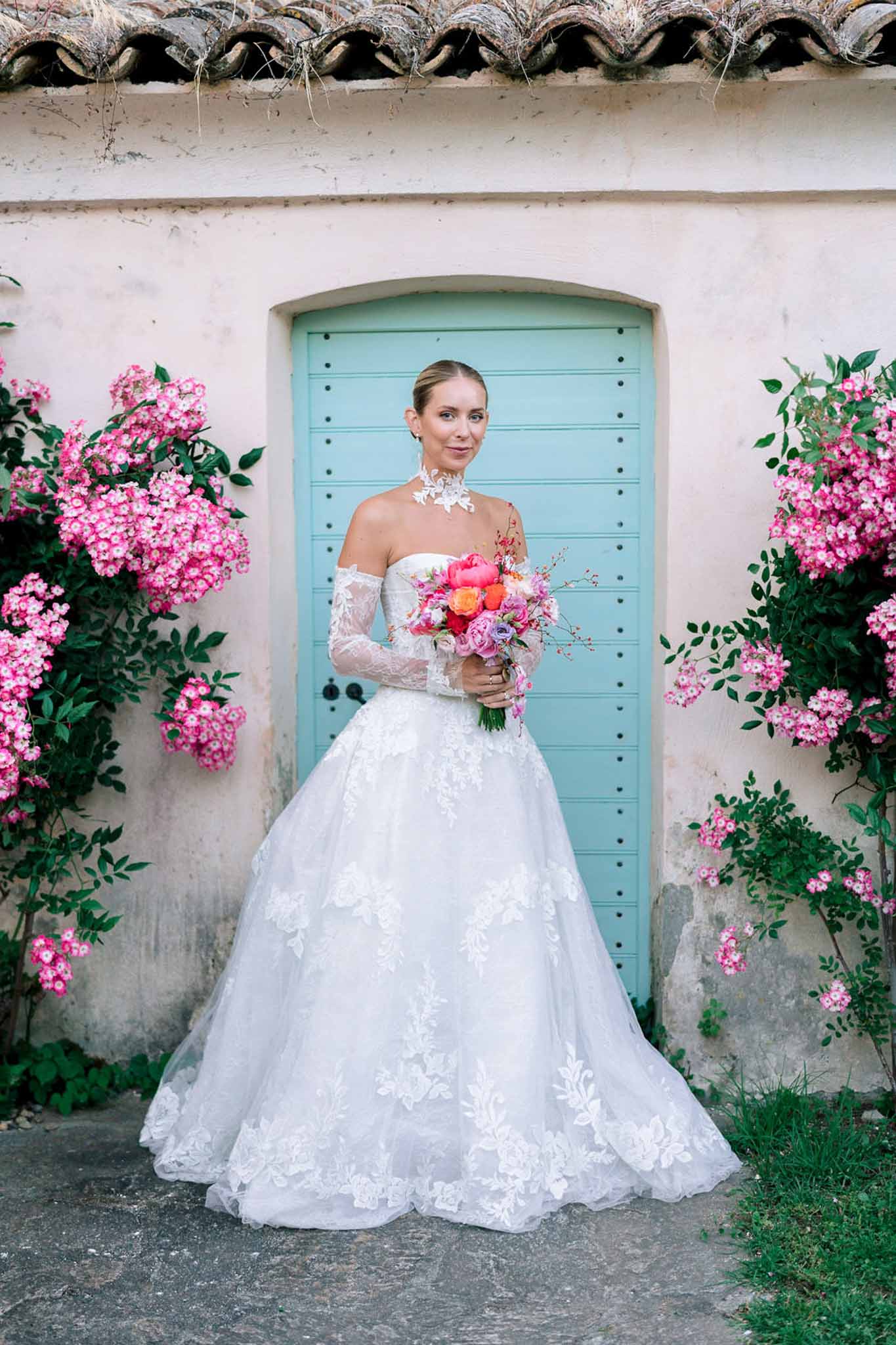 Bride in off-the-shoulder lace ball gown holding a vibrant pink peony and coral bouquet by a turquoise arched door