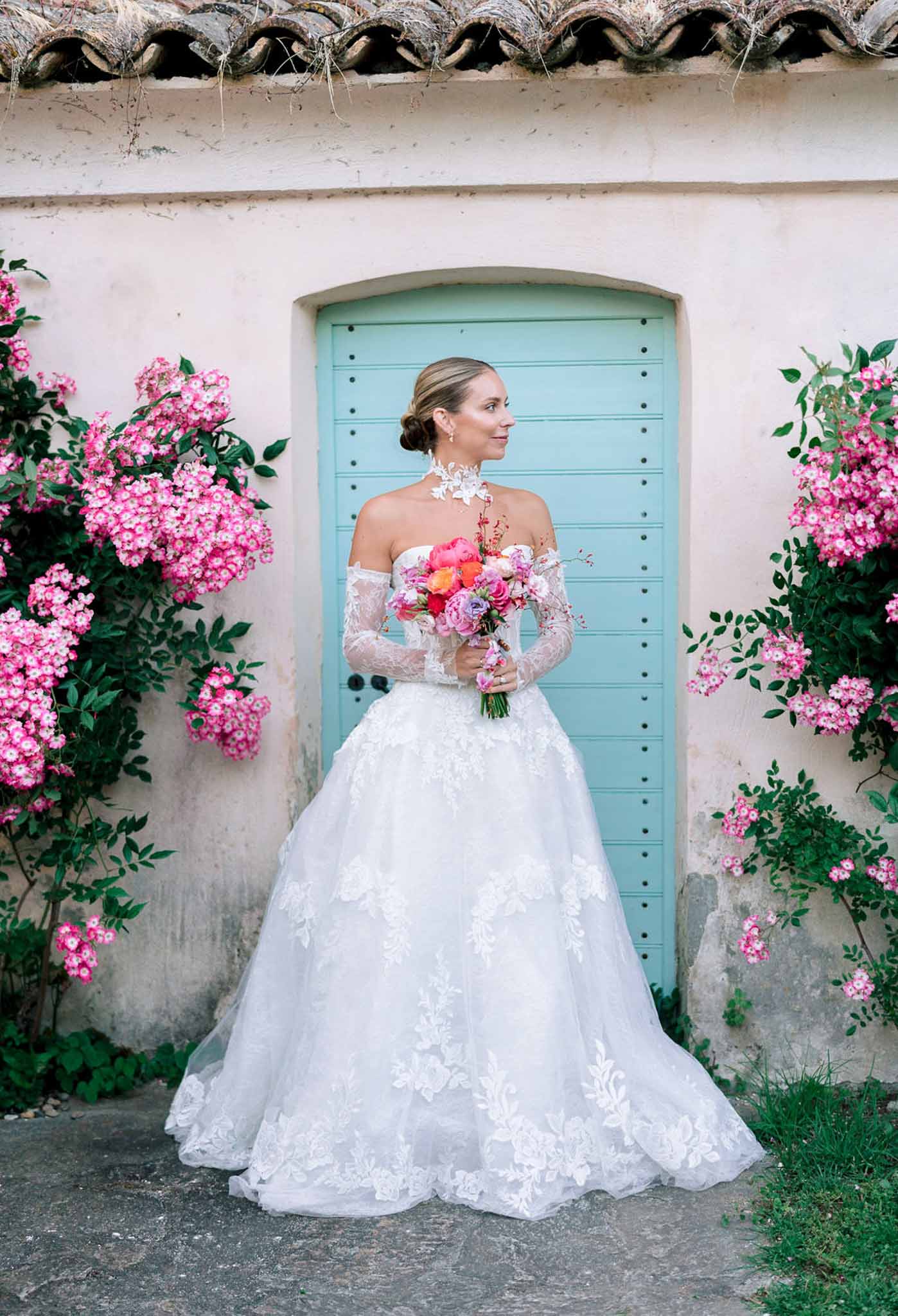 Bride in lace ballgown with hot pink peony bouquet before turquoise arched door with climbing roses