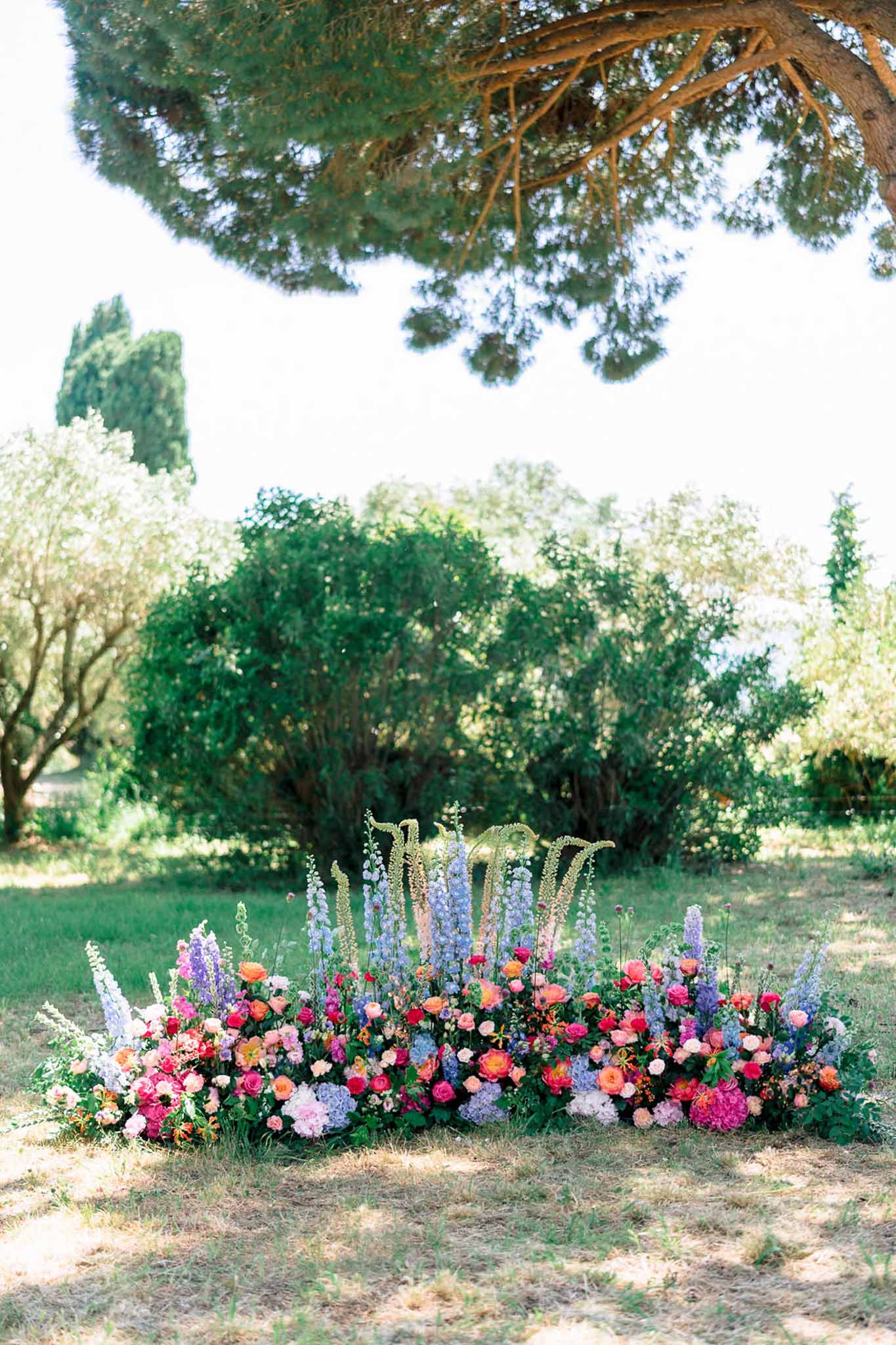 Ground-level ceremony flowers of hot pink roses, coral ranunculus, and periwinkle delphiniums with gold monogram