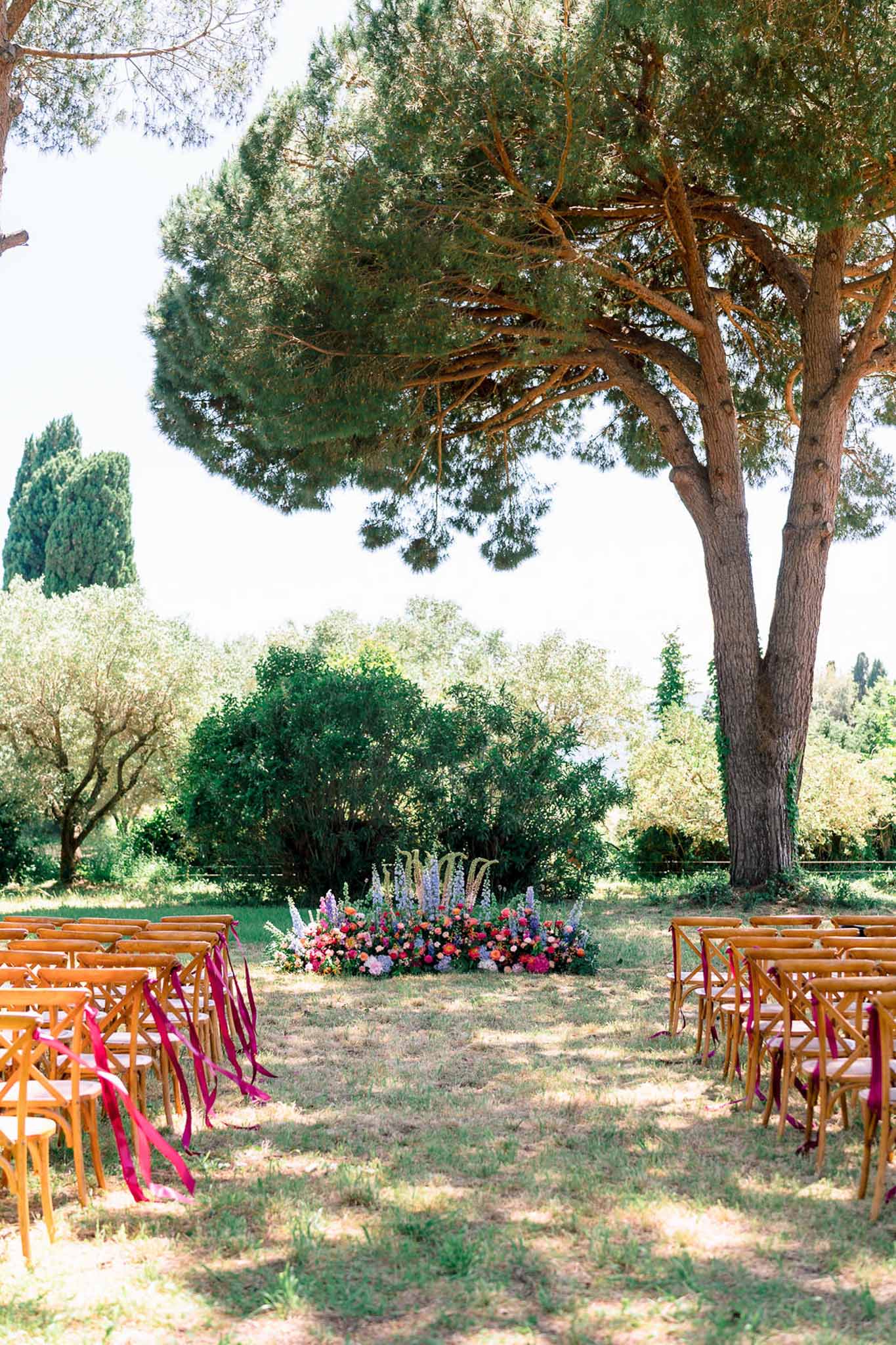 Ceremony aisle with fuchsia ribbon chairs and coral, pink, and lavender ground florals under umbrella pine