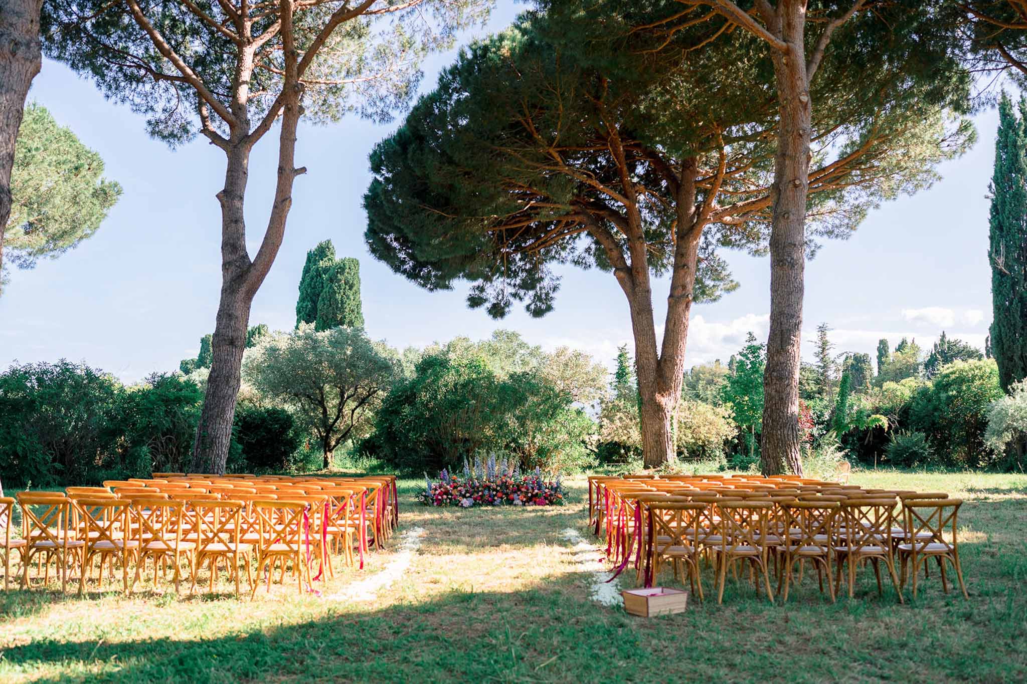 Cross-back chairs with fuchsia ribbons facing pink and purple ground florals under Mediterranean pines