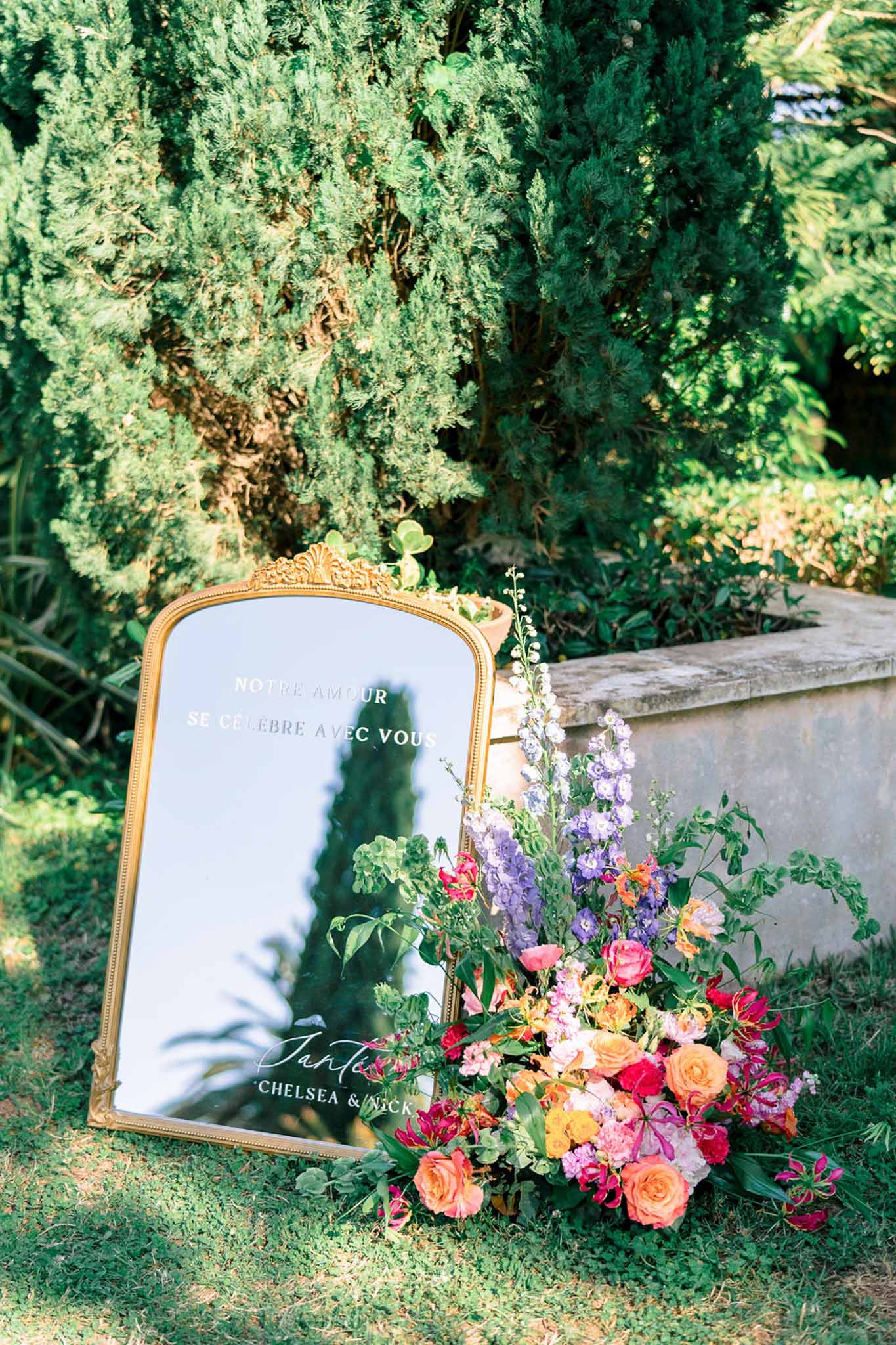 Gold mirror welcome sign with orange roses hot pink blooms lavender delphiniums and gloriosa lilies at garden ceremony