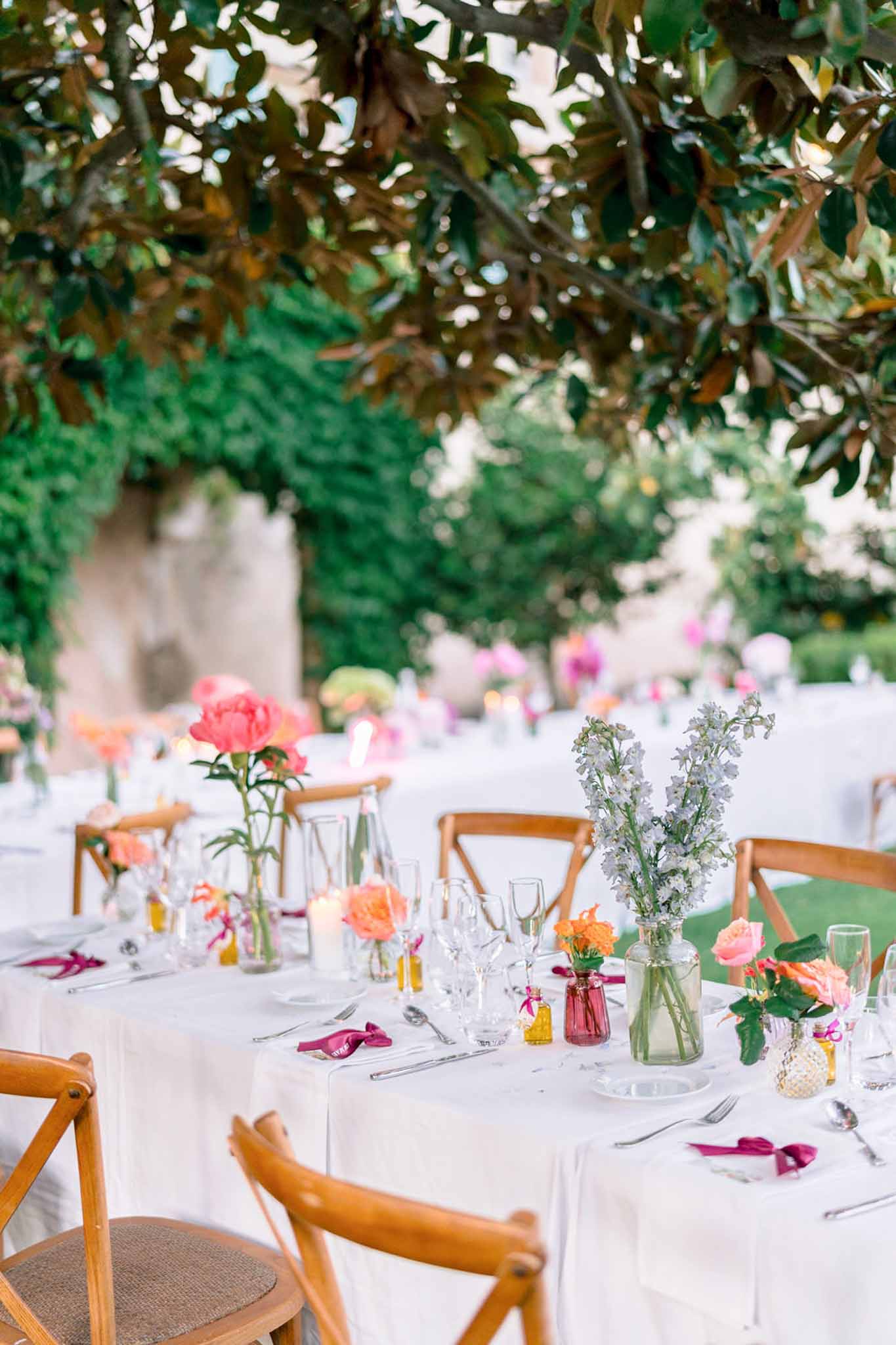 Outdoor reception tables with bud vases of coral peonies, peach roses, and blue delphinium on white linen